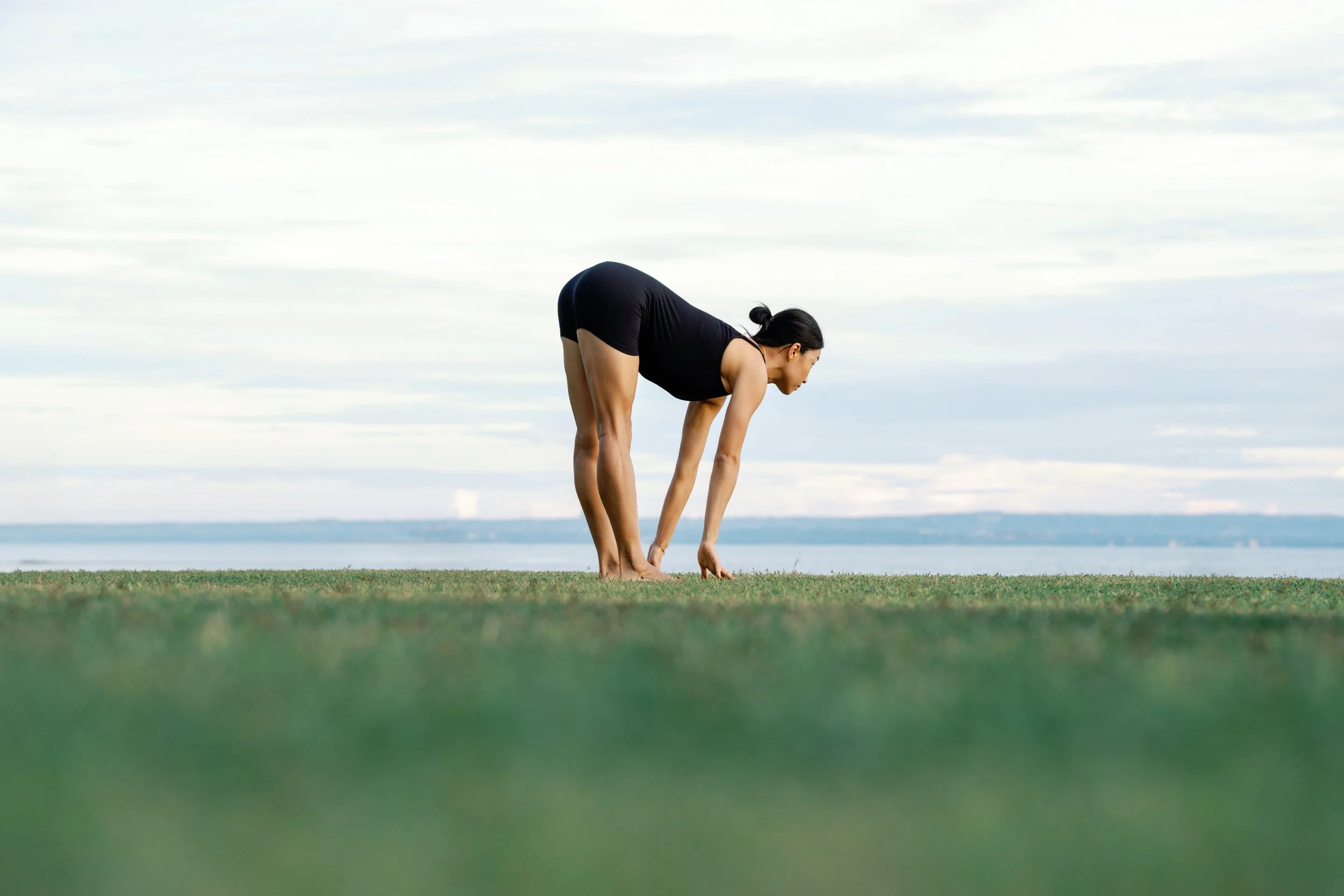 A woman in black athletic wear practicing yoga outdoors on grass near a body of water with land in the distance, under a partly cloudy sky.