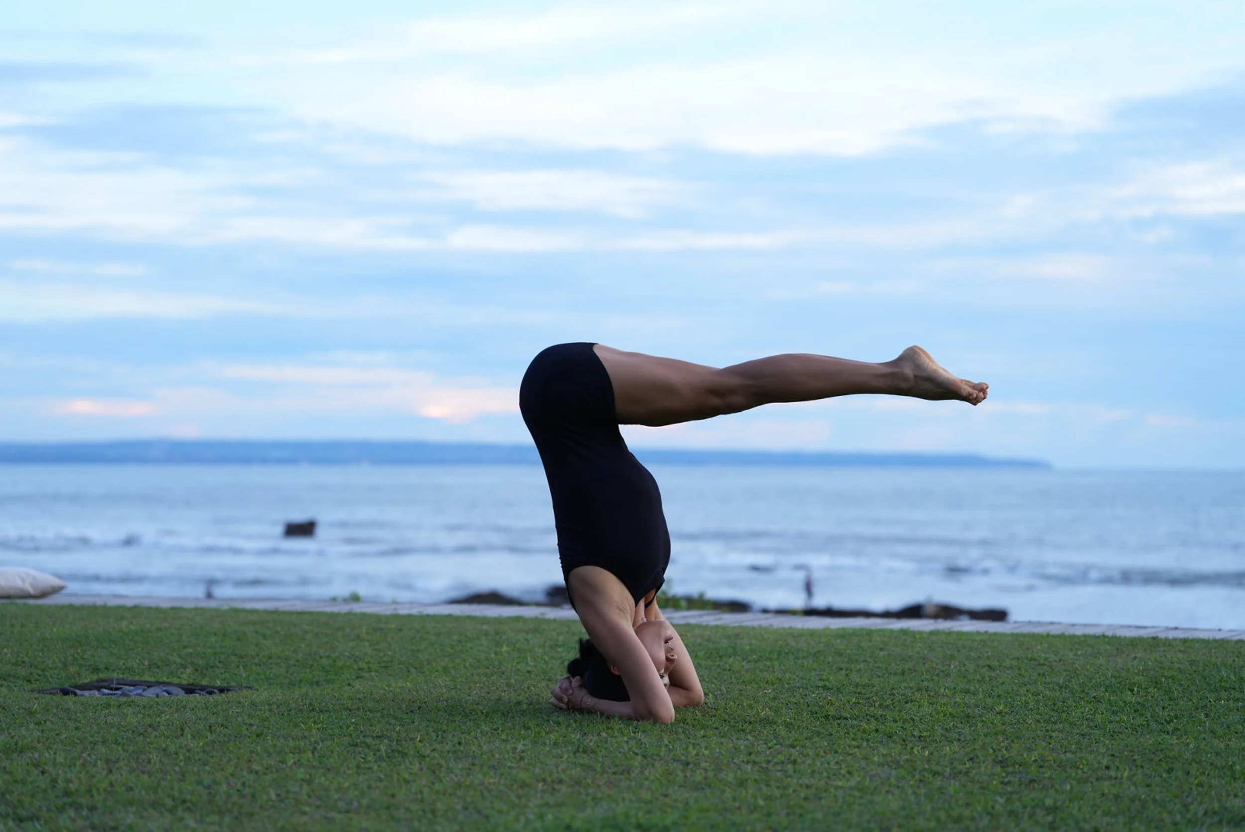 Person practicing yoga on grass near the ocean, performing a headstand with legs extended forward, in a peaceful outdoor setting.