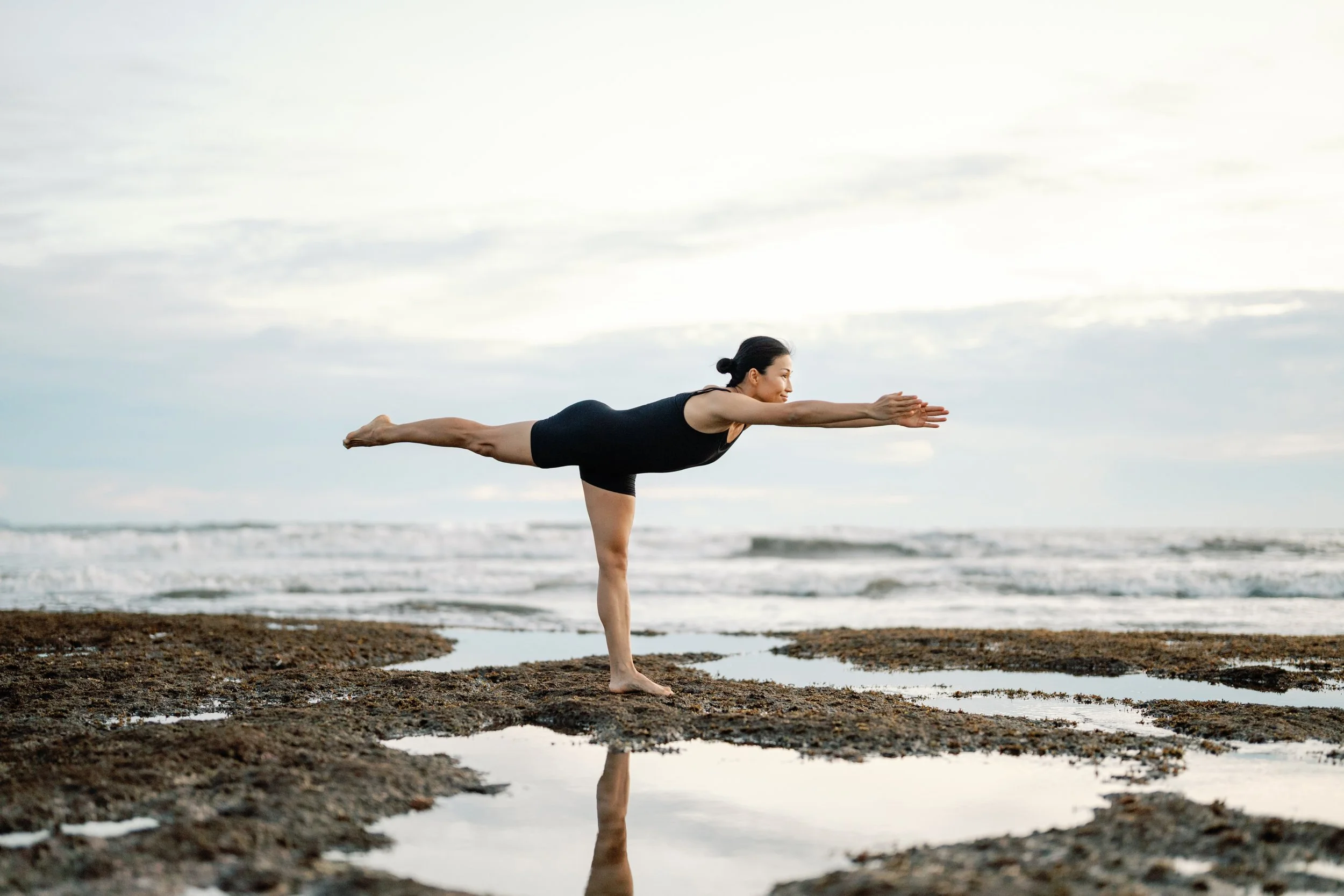 A woman practicing yoga on a beach, balancing on one leg while extending the other leg and her arms forward.
