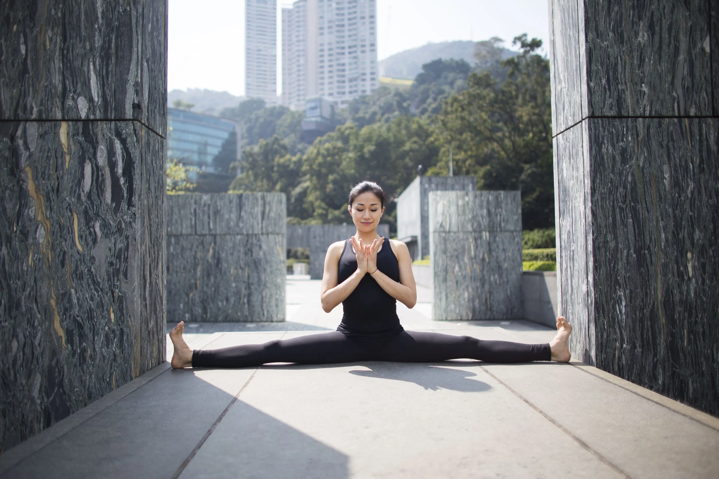Woman in black yoga outfit doing a seated split yoga pose outdoors with city buildings and trees in the background.