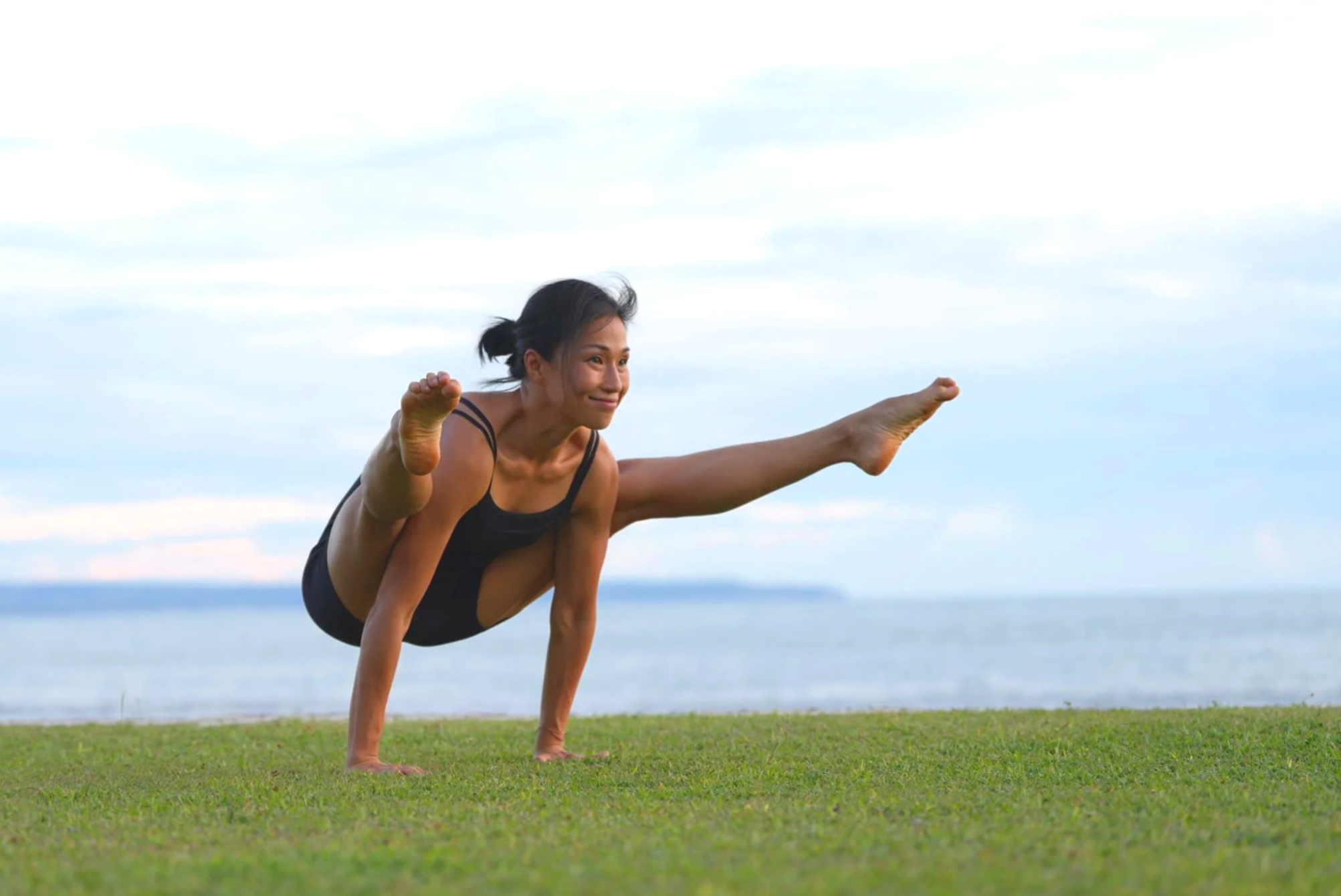 Woman practicing yoga outdoors on grassy field with ocean and cloudy sky background, balancing on one hand with one leg extended sideways and the other knee bent.