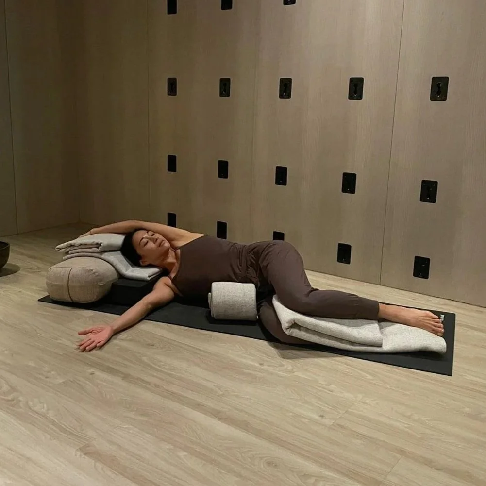 A woman lying on an eco-friendly yoga mat, resting her head on a pillow with a bolster and a folded towel, in a wellness or yoga studio.