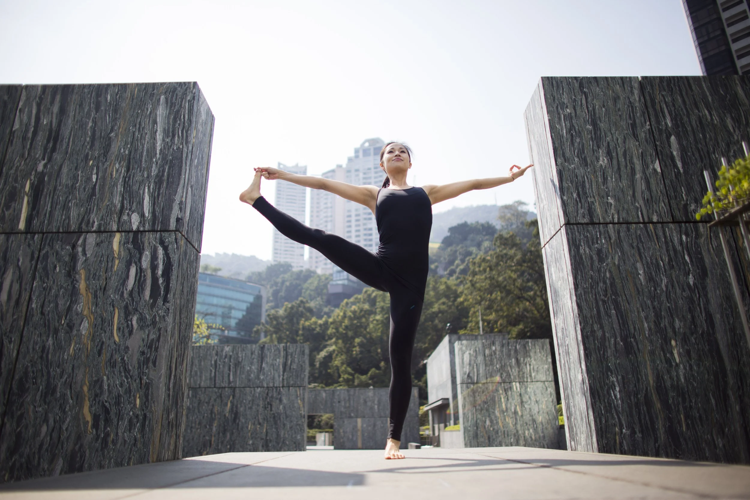 A woman in black yoga attire performs a standing split pose outdoors between two large stone walls with a cityscape and mountains in the background.