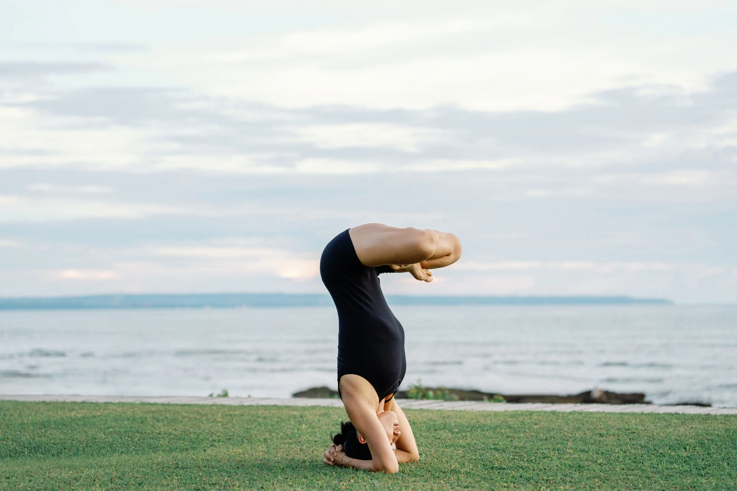 Person performing a yoga headstand on grass near a body of water with cloudy sky in the background.