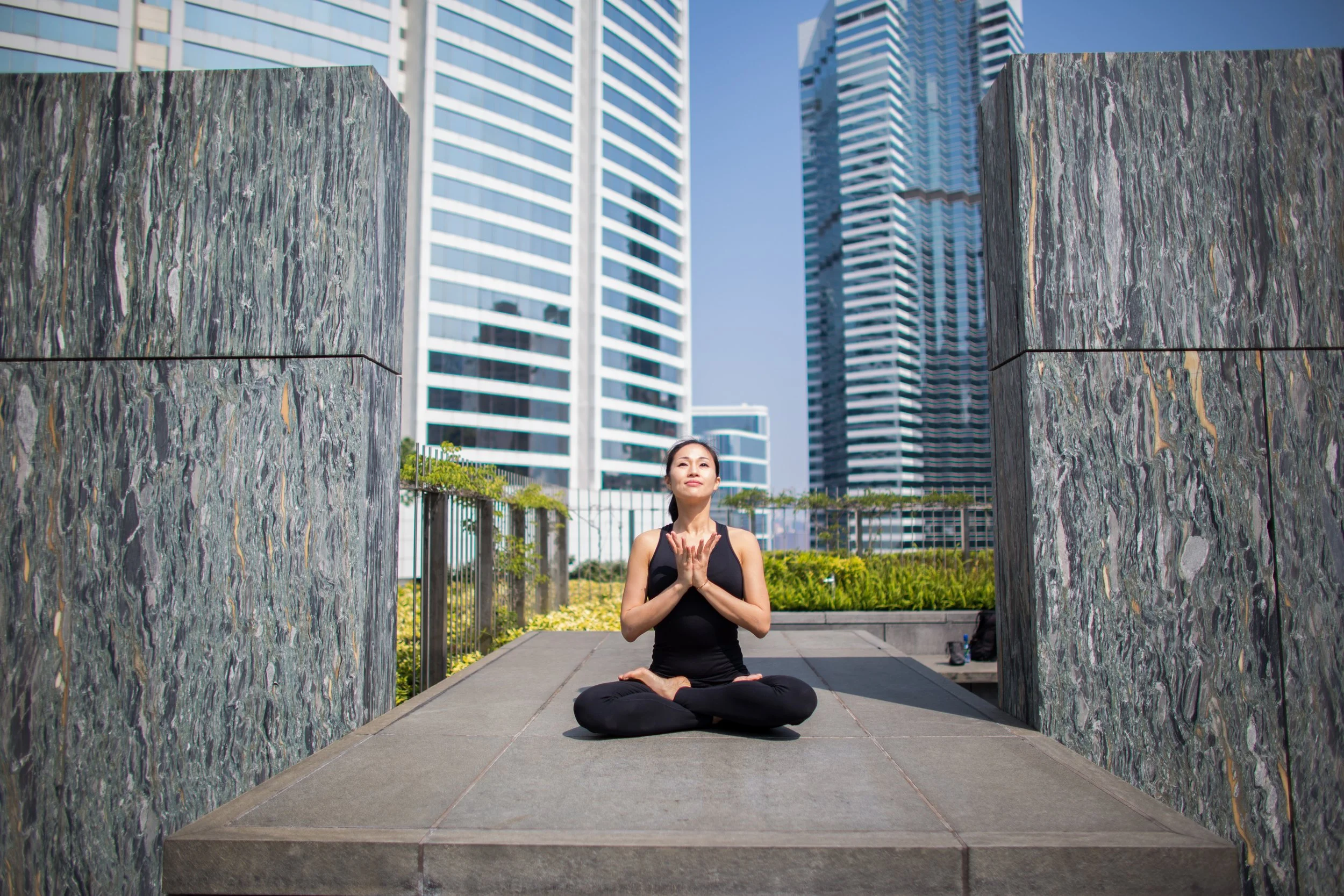 A woman practicing yoga outdoors on a city rooftop, sitting cross-legged with hands in prayer position against tall modern skyscrapers and a clear blue sky.