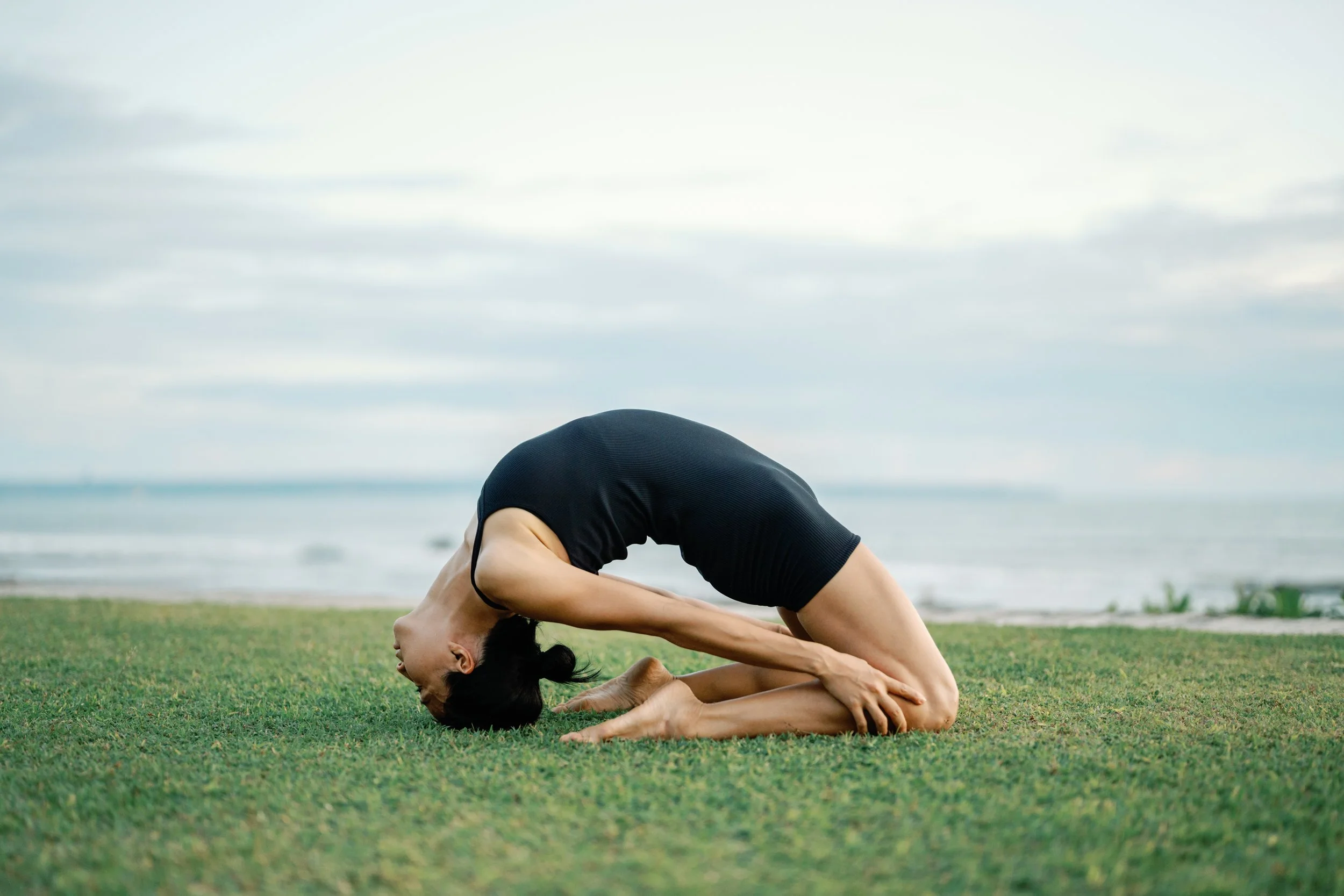 A woman practicing yoga on the grass near the beach, performing a forward bend with her head on the ground, wearing a black top and black pants.