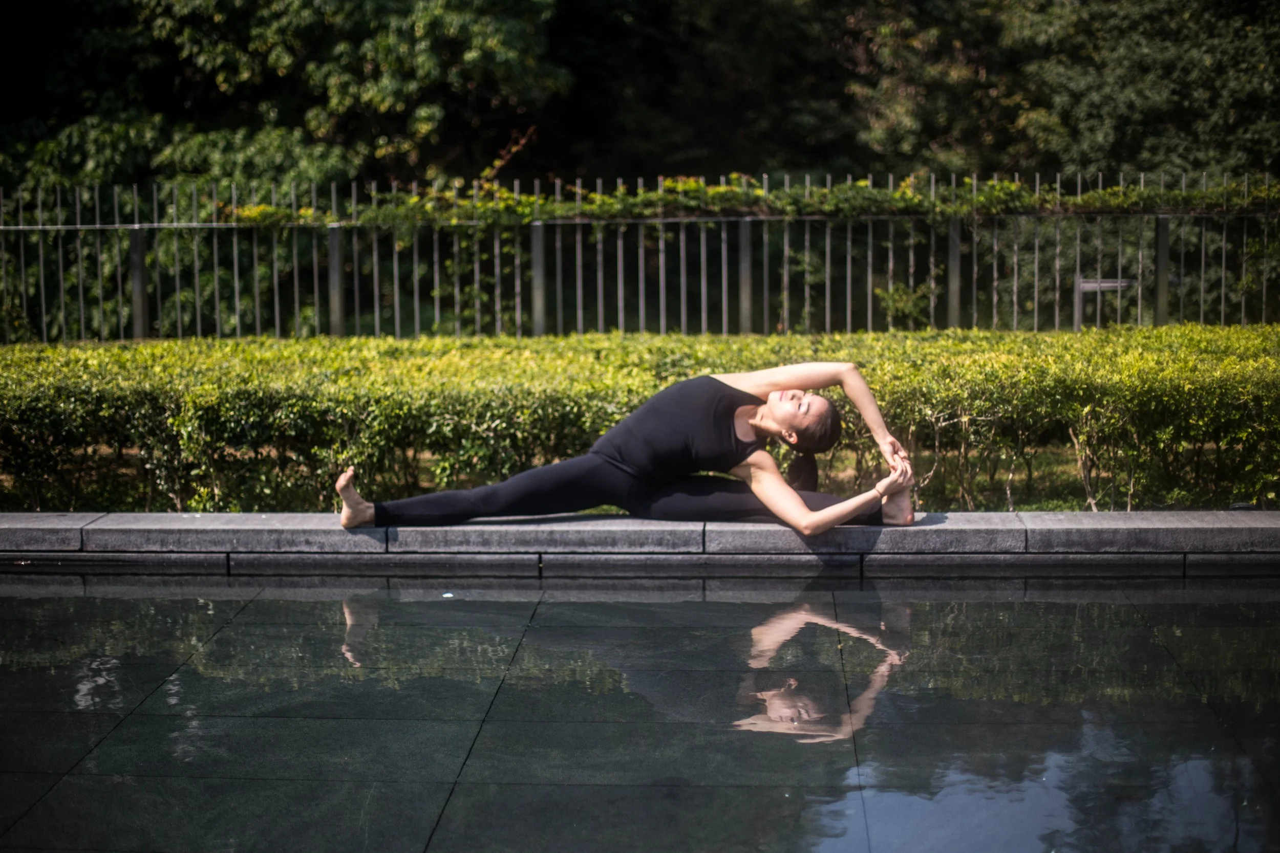 A woman practicing yoga outdoors beside a reflecting pool, performing a side stretch with one arm reaching over her head and holding her foot, dressed in black activewear.