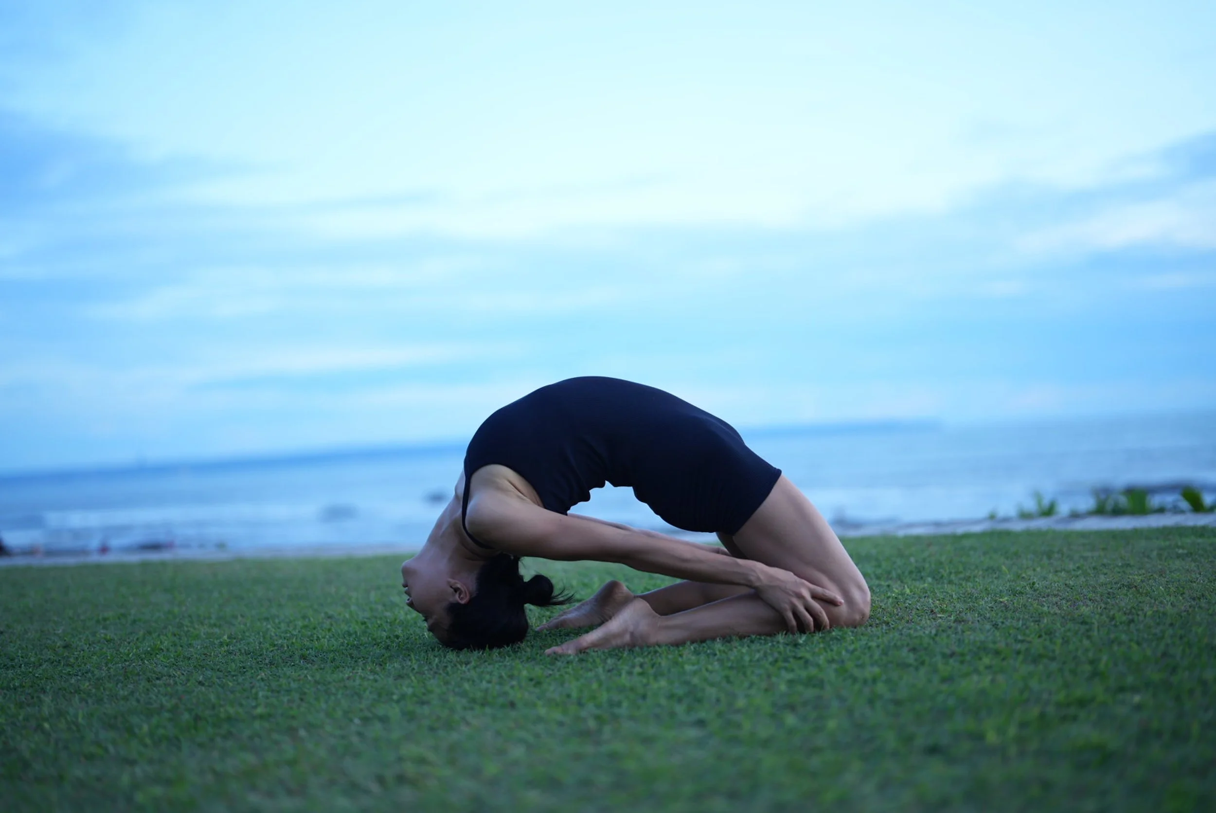 Person practicing yoga on grass near the beach, performing a forward bend with their hands holding their feet.