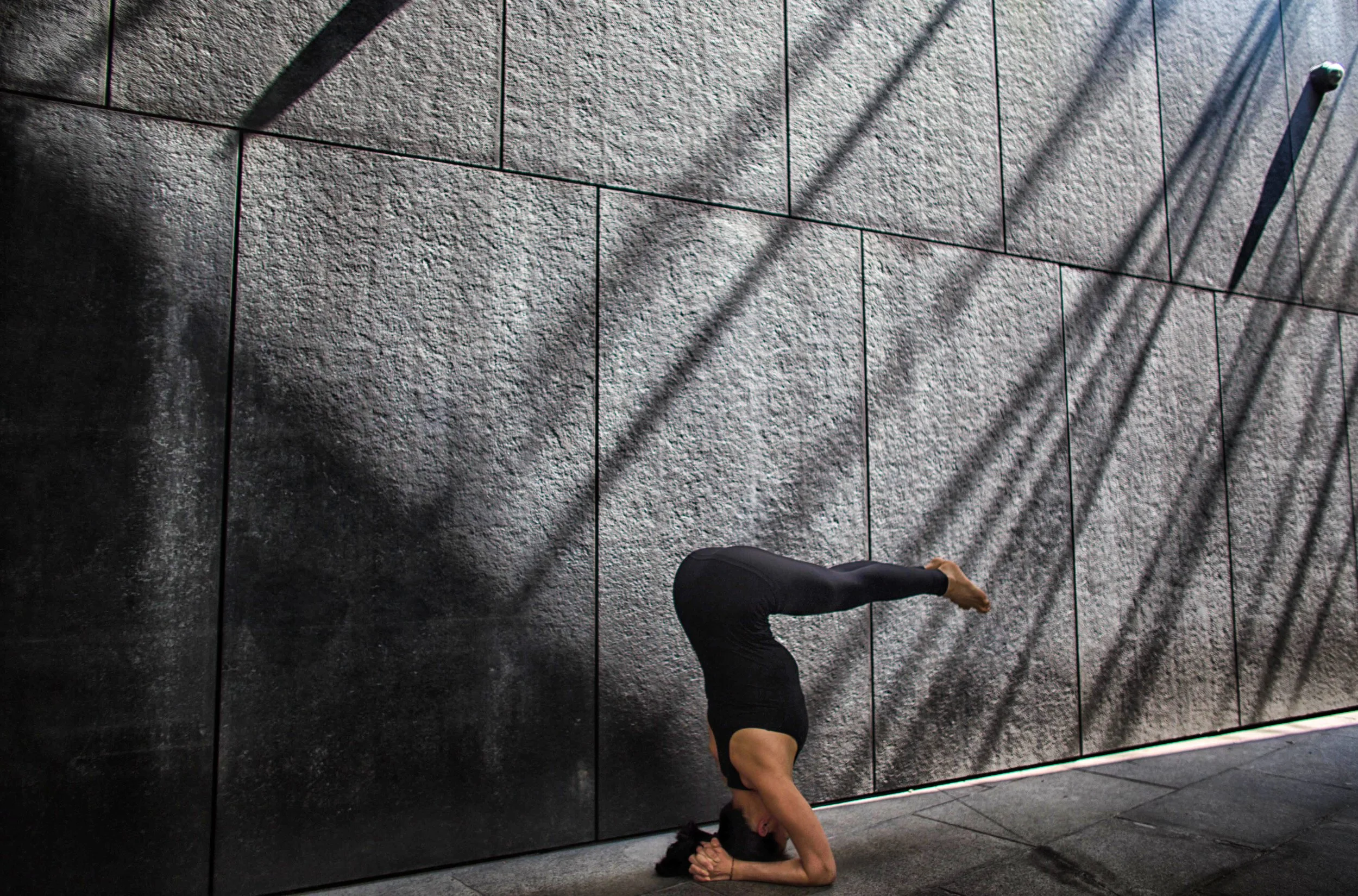 A person doing a headstand against a textured gray wall casting shadow patterns.