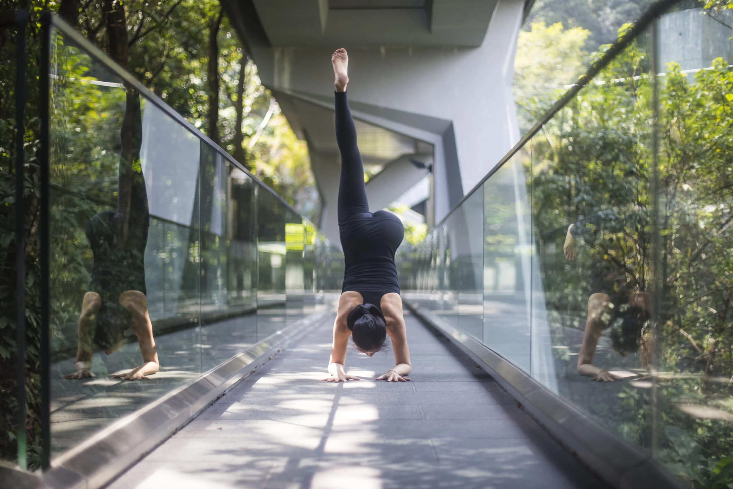 A woman performs a handstand in a yoga pose on a glass walkway surrounded by greenery.