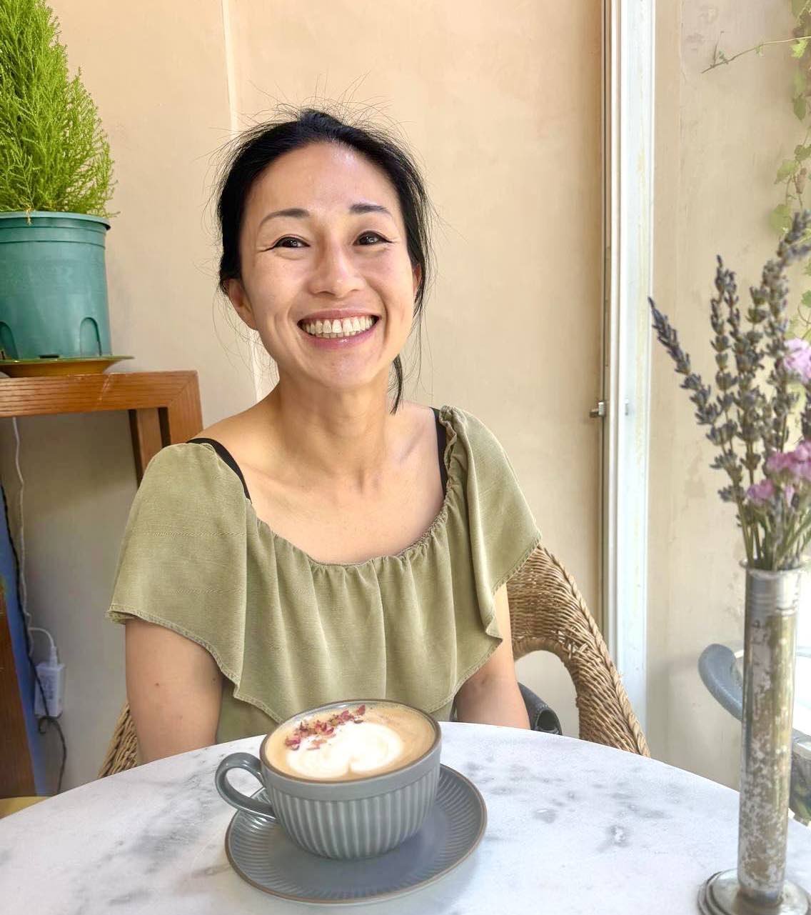 A woman smiling at a cafe, sitting at a table with a cup of coffee topped with whipped cream and pink flowers, with a vase of purple flowers nearby.