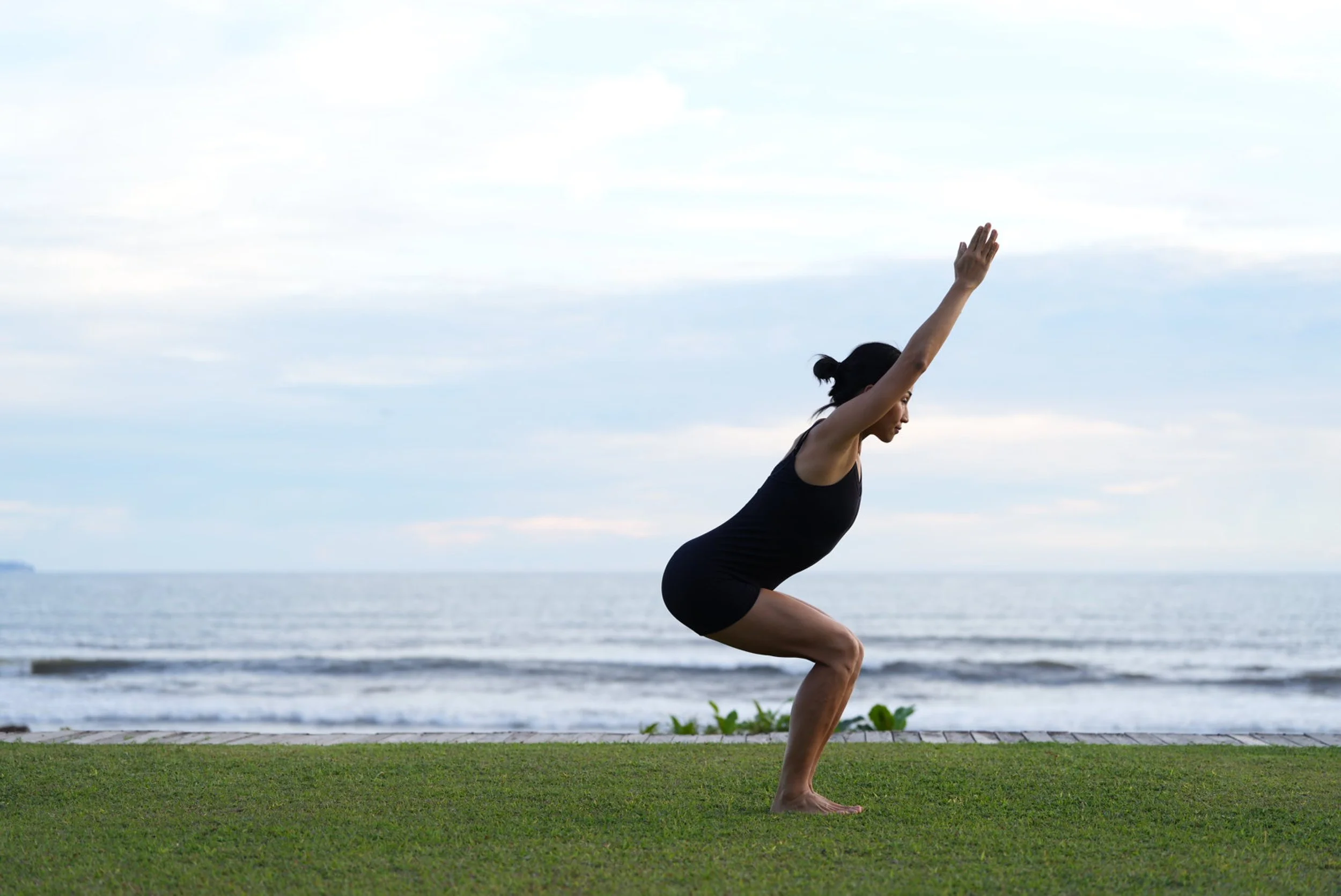 A woman practicing yoga outdoors on a grassy area near the beach, performing a squat with her arms raised overhead, facing the ocean.