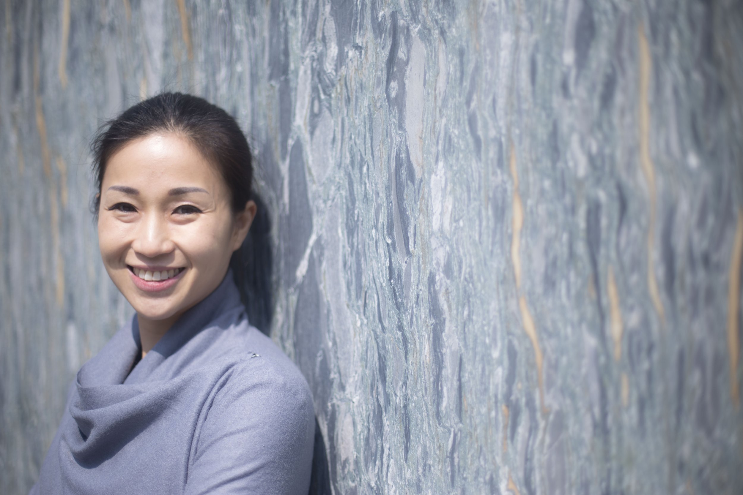 Smiling woman with dark hair, wearing a gray top, leaning against a textured blue-gray wall outdoors.