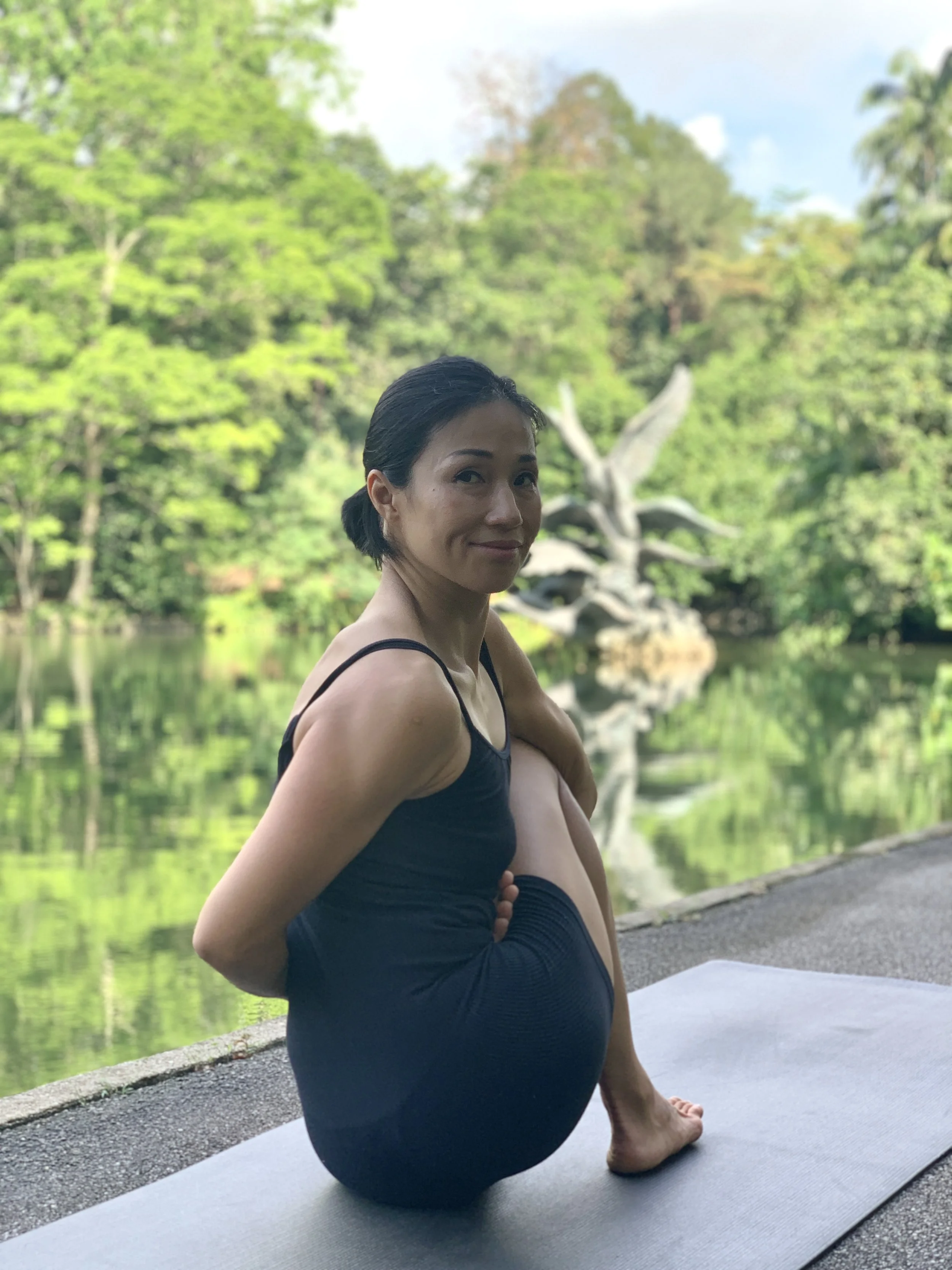 A woman practicing yoga outdoors near a pond, sitting with her legs crossed and arms behind her back, with a serene natural background of trees and a sculpture in the distance.