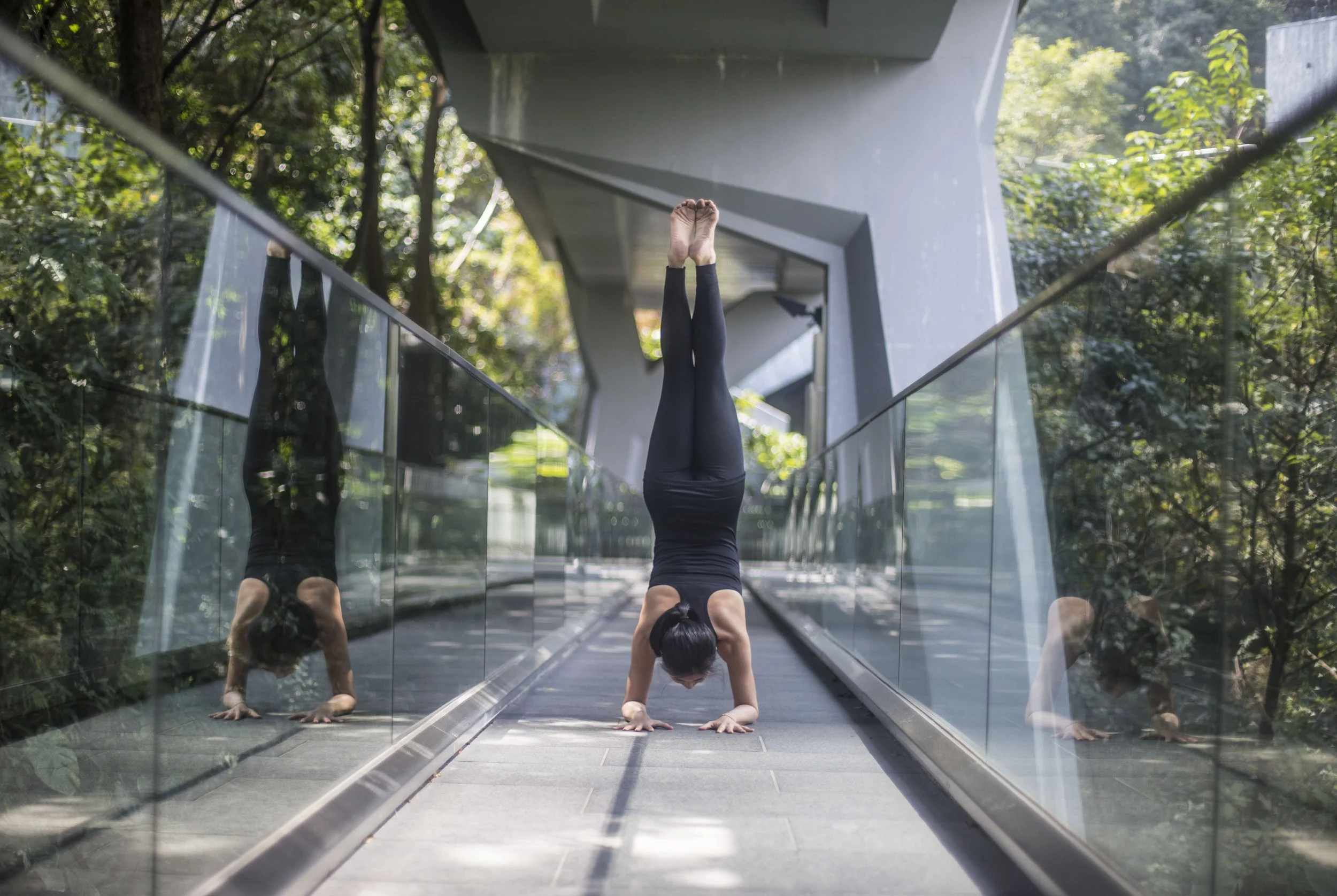 A woman doing a handstand in a yoga pose outdoors on a bridge with glass railings and a modern architectural structure overhead. She is wearing black athletic clothing and the surroundings include green foliage.