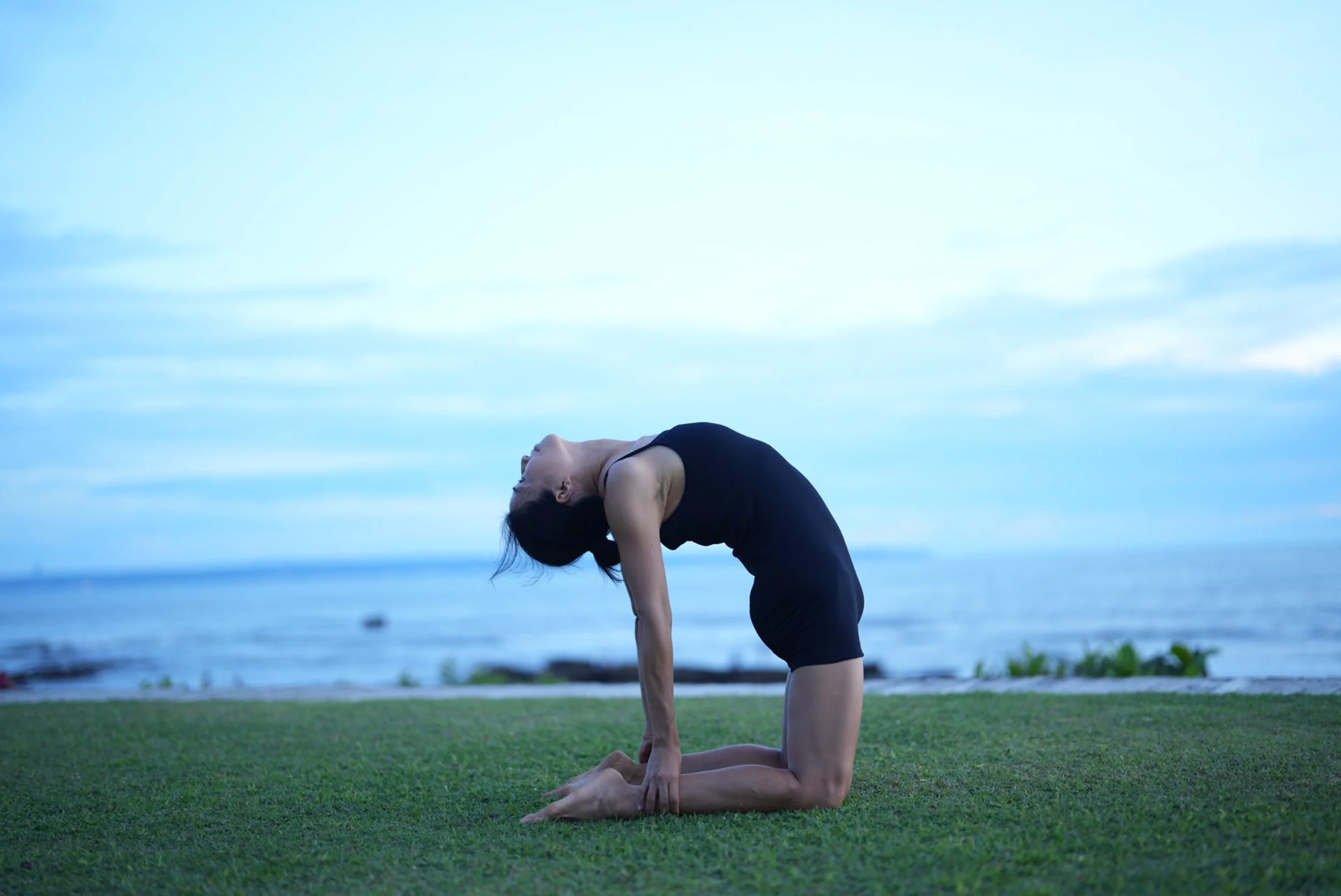 Woman practicing yoga near the ocean, kneeling on grass with a dramatic backbend.