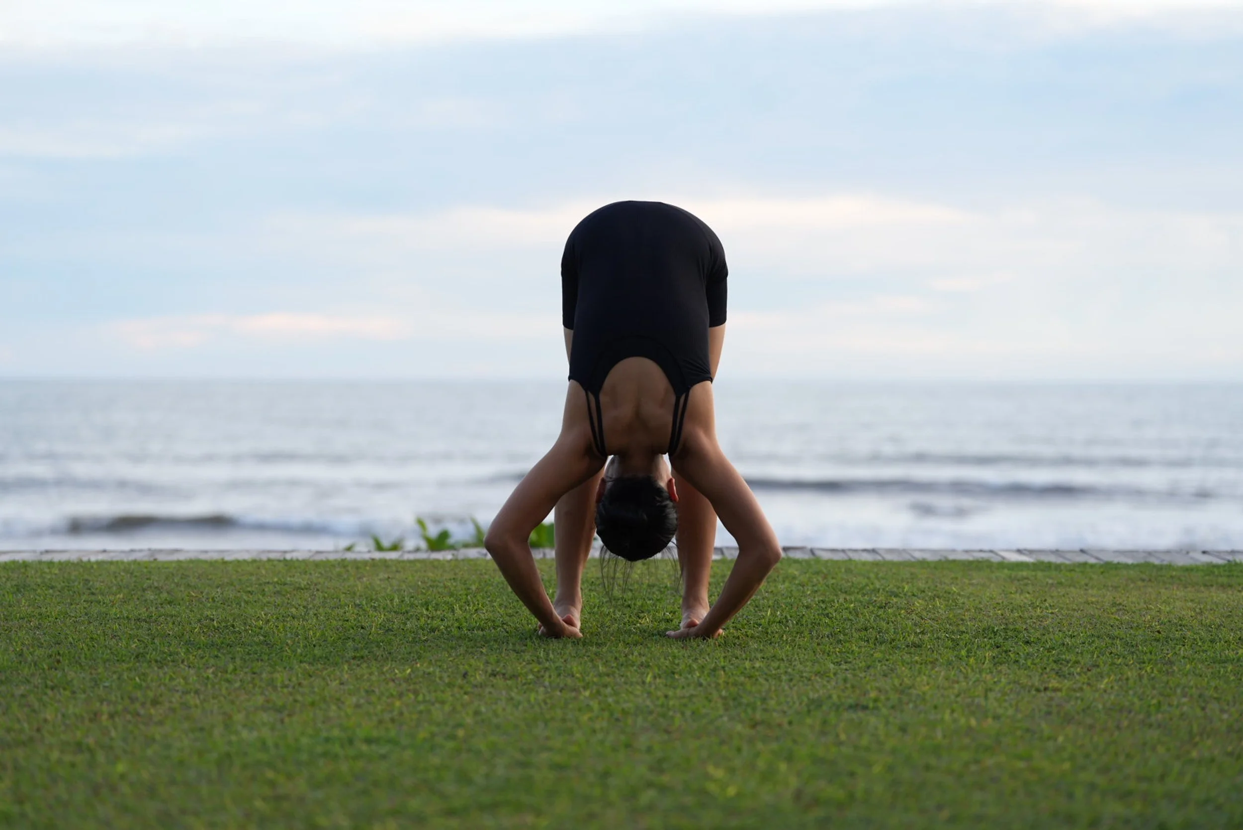 A woman doing a downward dog yoga pose on a grassy area near the beach, facing towards the ocean with a cloudy sky above.
