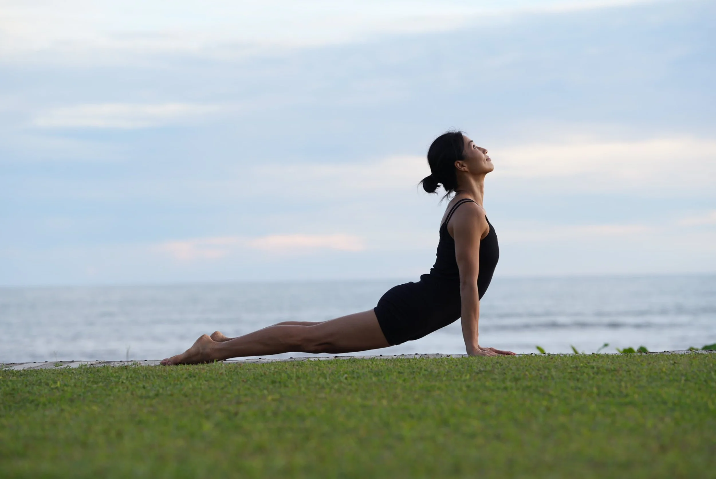 A woman practicing yoga outdoors on grass near the ocean, performing a reverse plank pose with her head tilted back and eyes closed.