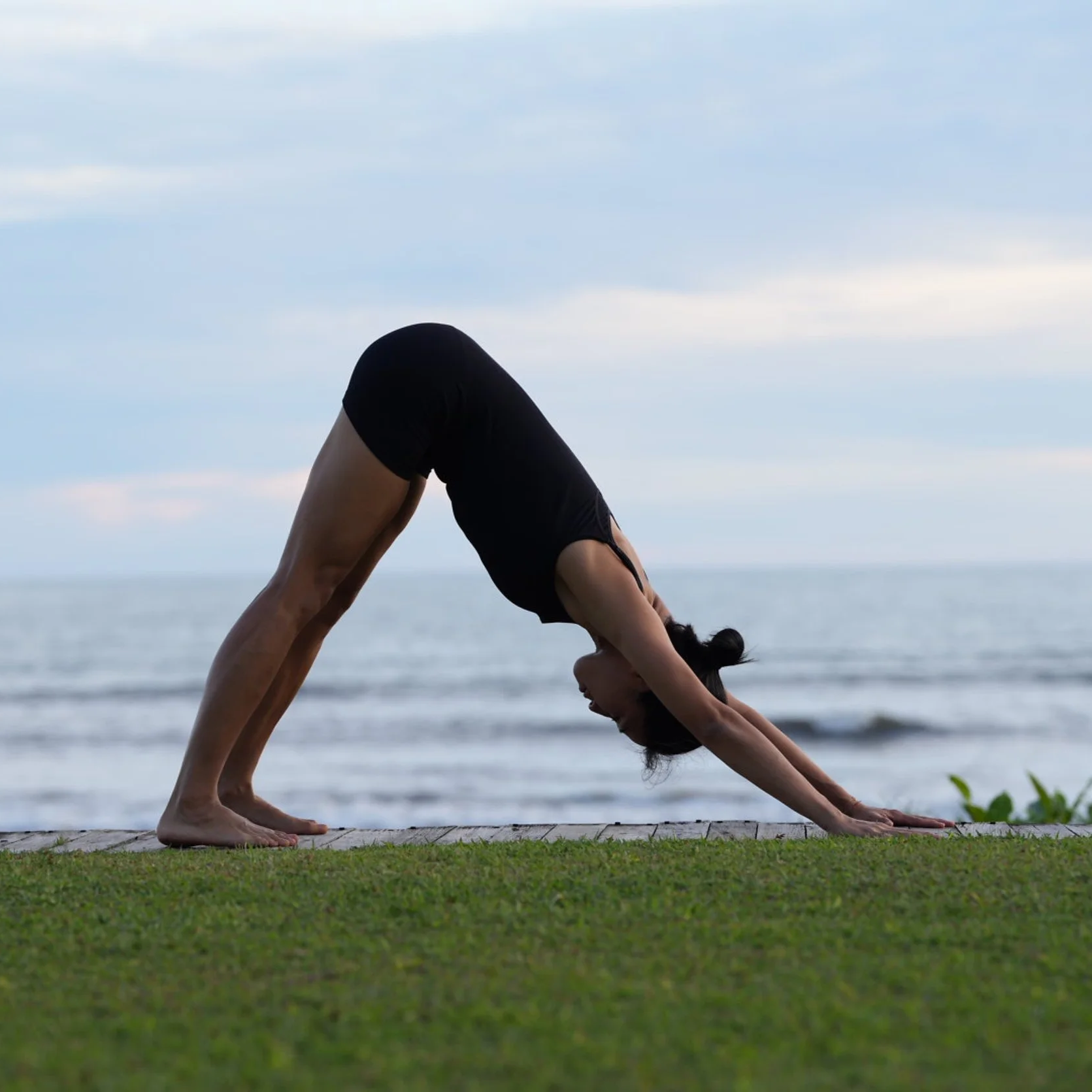 A woman practicing yoga outdoors on a grassy area near the beach, performing the downward dog pose with her hands and feet on the ground, against a backdrop of ocean and cloudy sky.