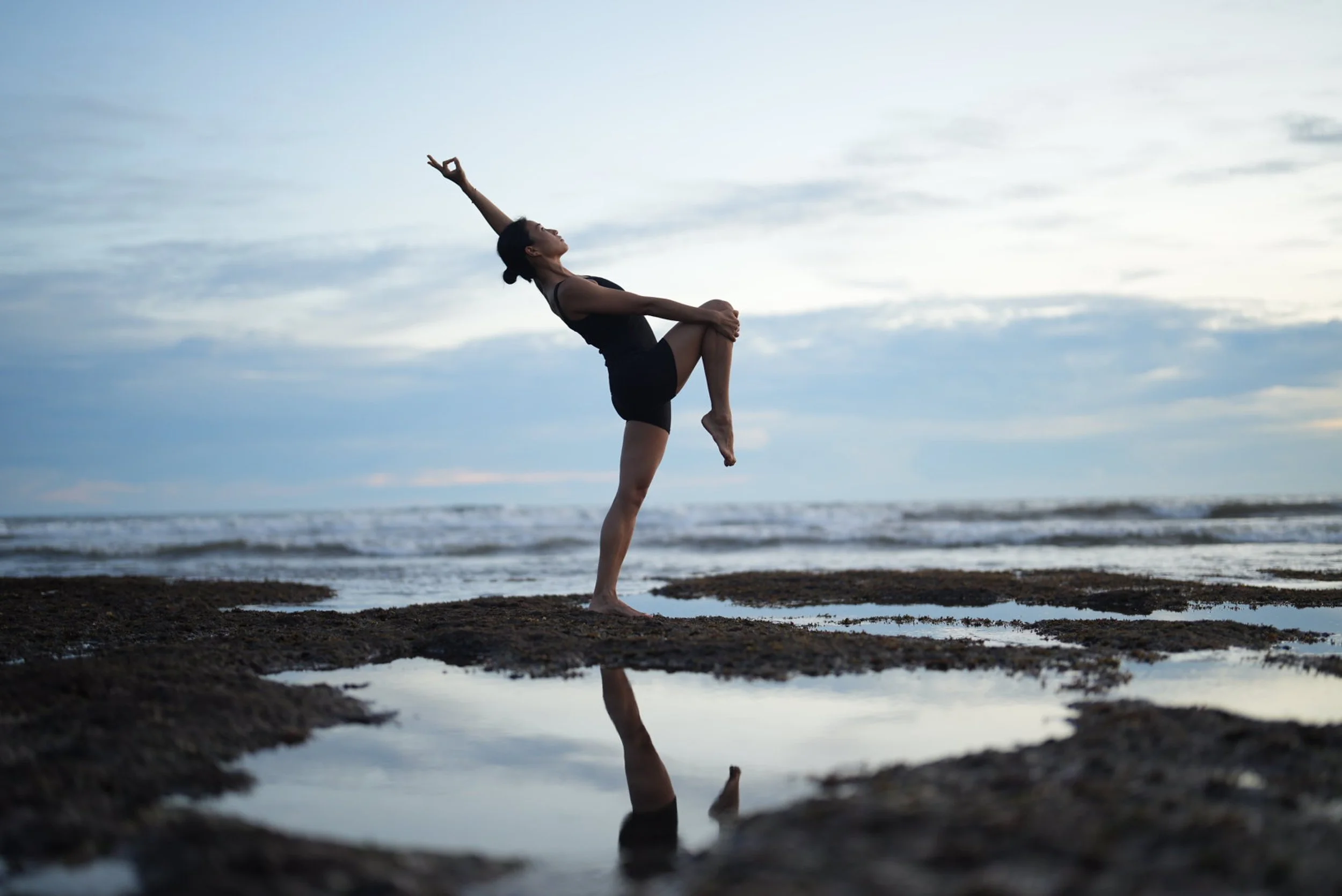 A woman performing a yoga pose on a rocky beach during sunset with the ocean and cloudy sky in the background.