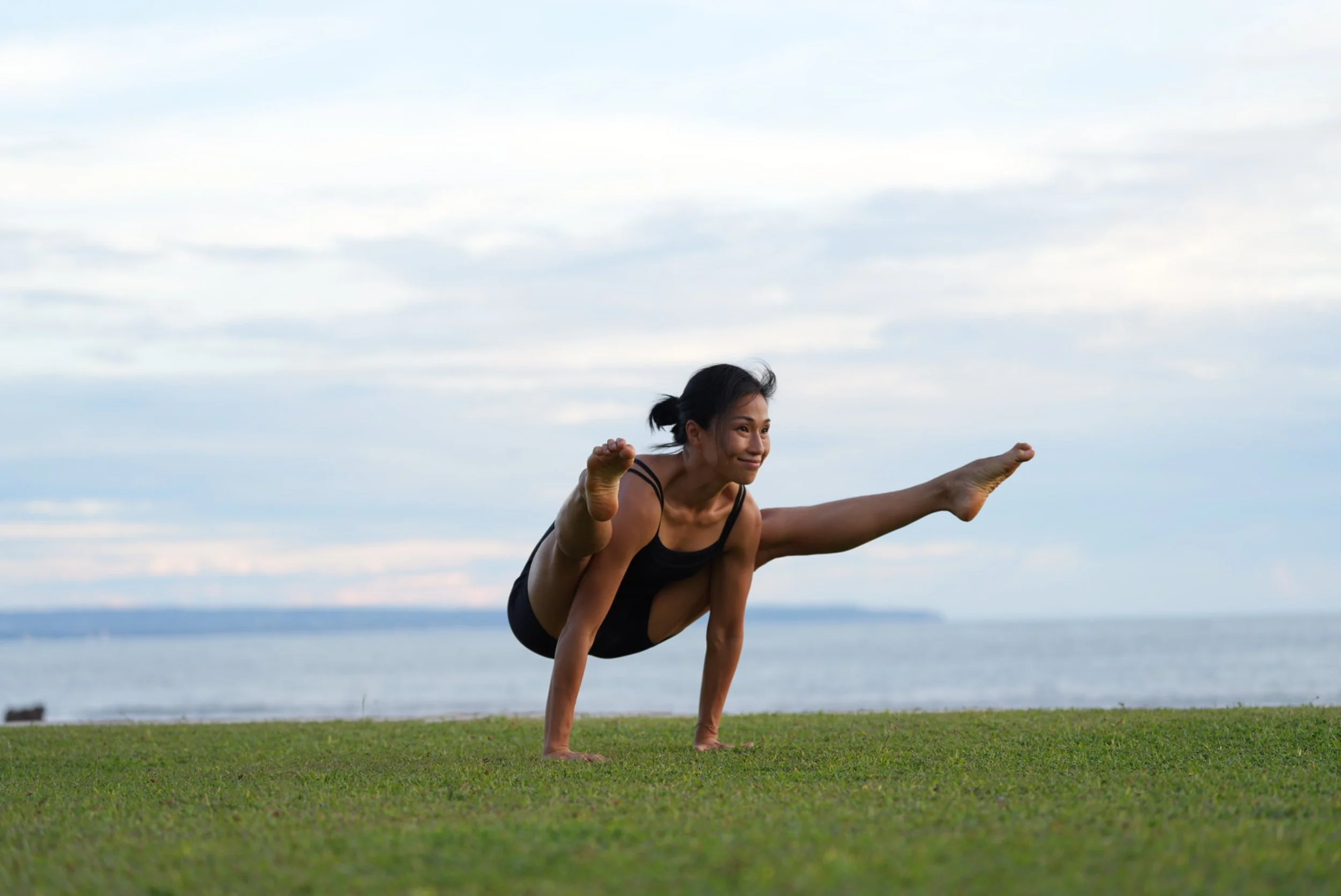 Woman practicing yoga or calisthenics outdoors on grass near water, balancing on one hand with her legs extended and arms stretched.
