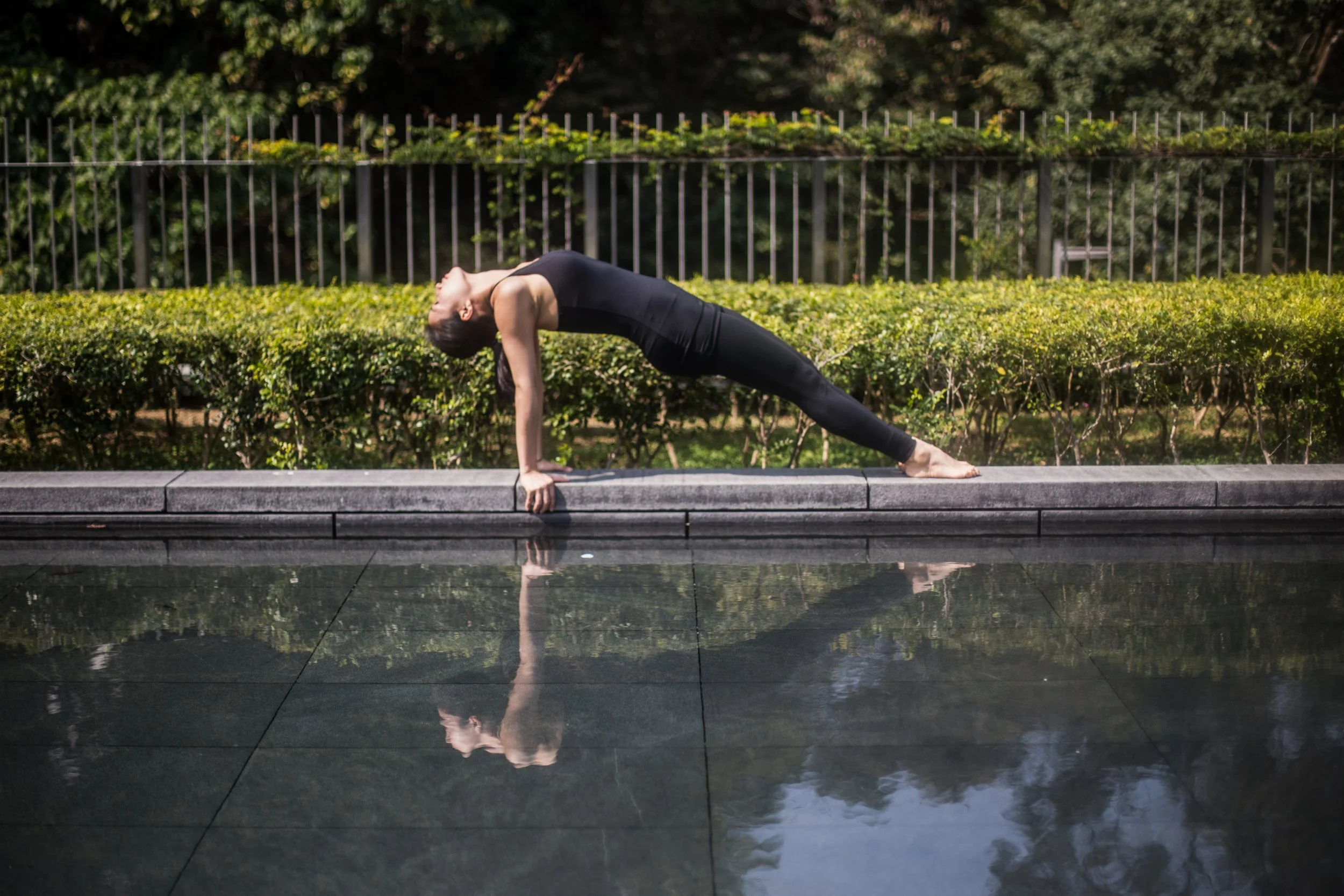 A woman practicing yoga outdoors beside a reflective pool, doing a downward dog pose with her head hanging down, in a peaceful garden setting.