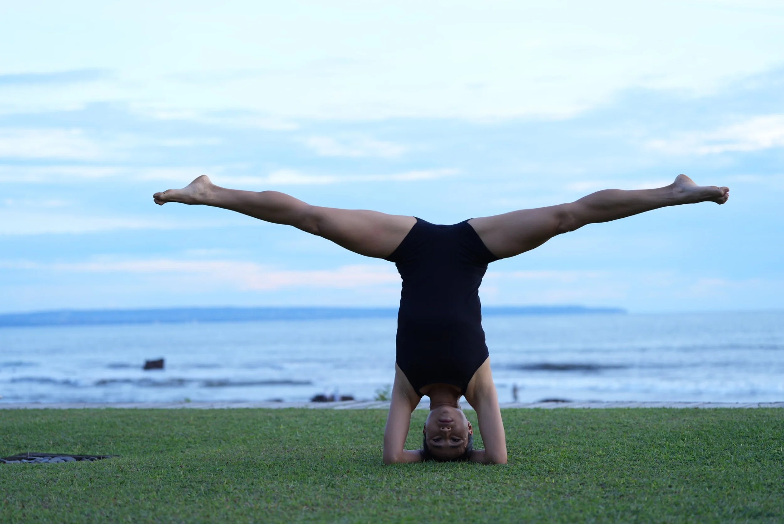 Person performing handstand on green grass near ocean with cloudy sky in the background.