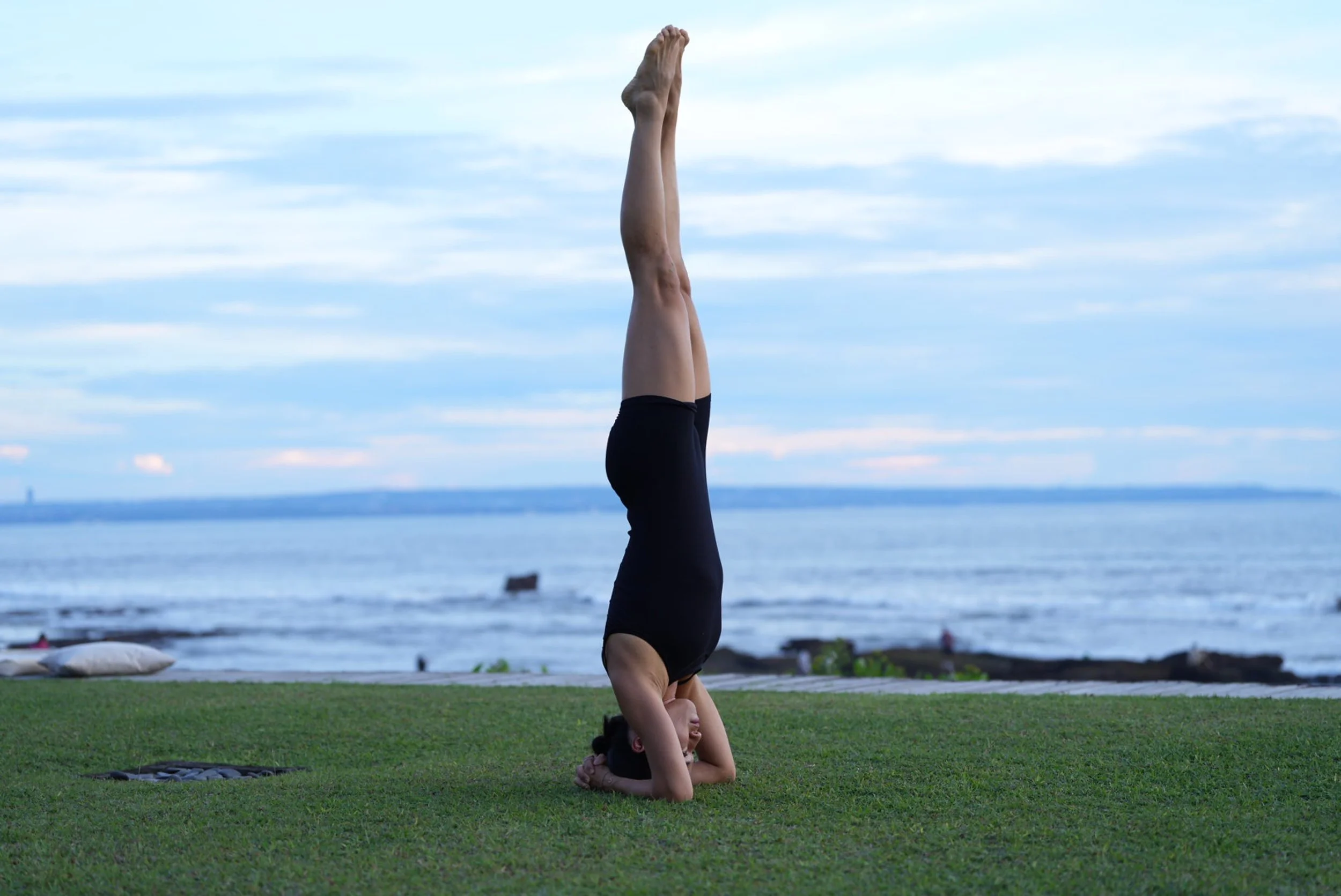 A woman practicing yoga outdoors on grass near the ocean, performing a headstand with her arms supporting her head and neck, against a backdrop of blue sky and water.