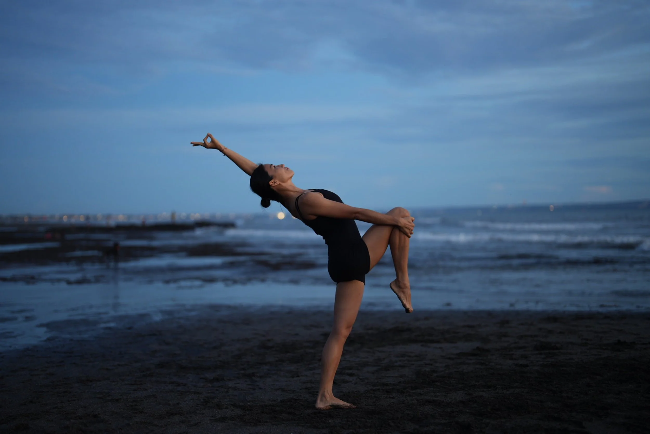 A woman practicing yoga on a dark sandy beach near the ocean at dusk, wearing a black athletic outfit, balancing on one leg with one arm extended upwards and the other holding her knee.