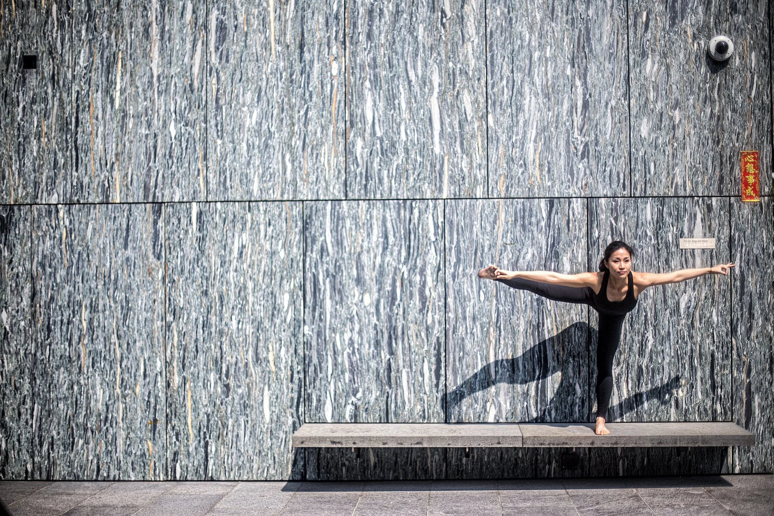 A woman practicing yoga, balancing on one leg with her other leg extended to the side, against a textured gray stone wall.