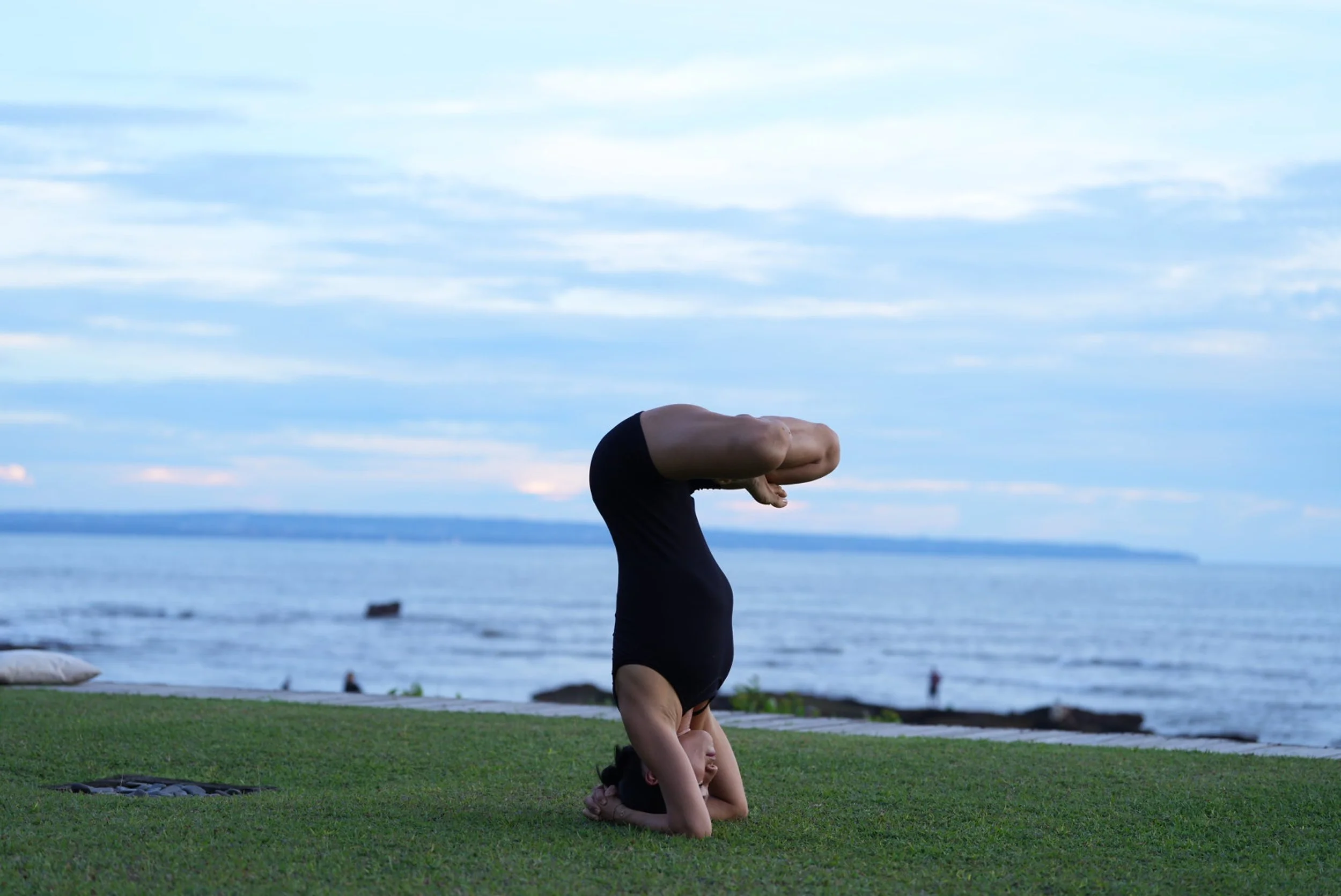Person practicing a headstand yoga pose on the grass near a body of water with a cloudy sky.