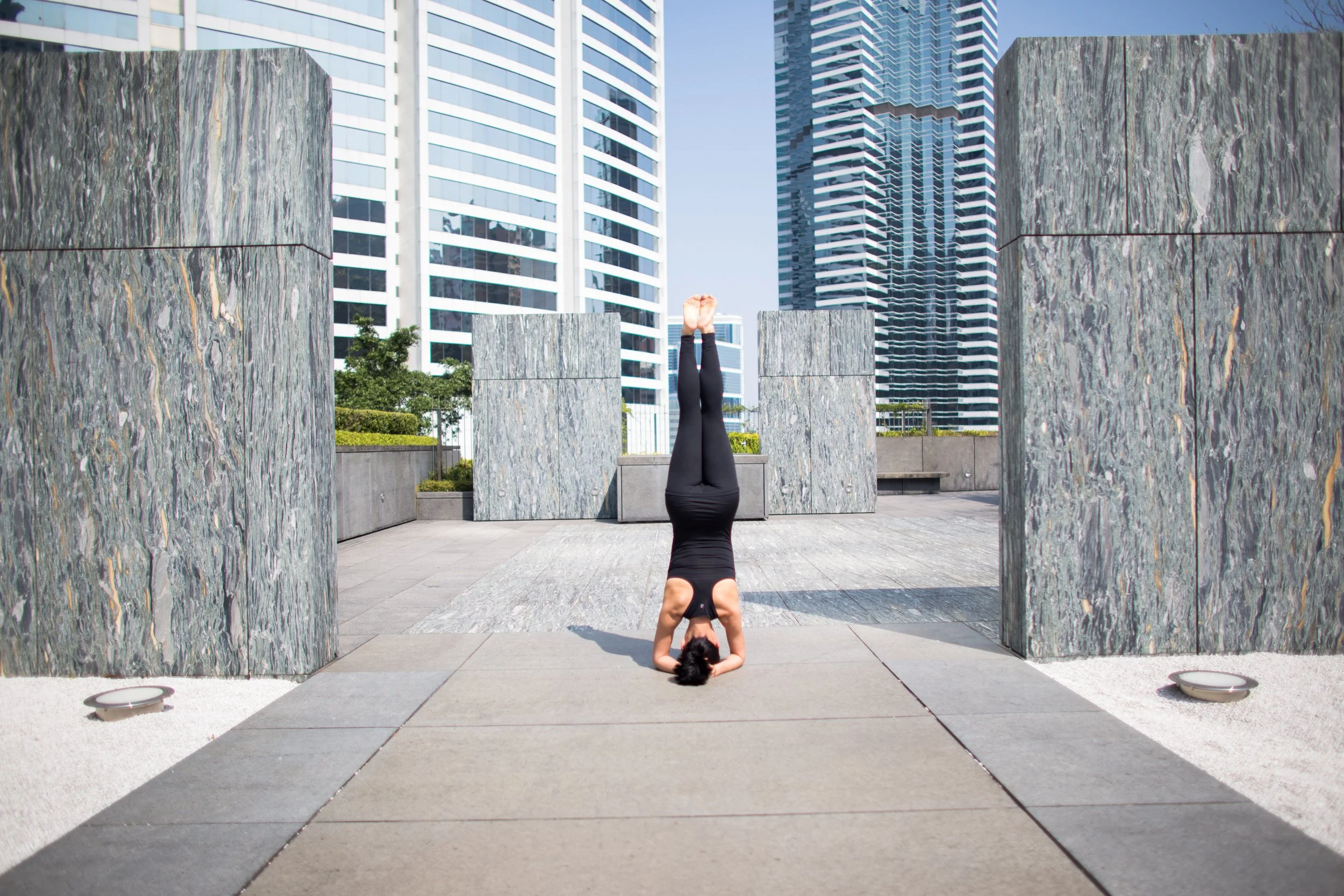A woman practices headstanding yoga pose outdoors in an urban setting with modern skyscrapers in the background and stone structures around her.