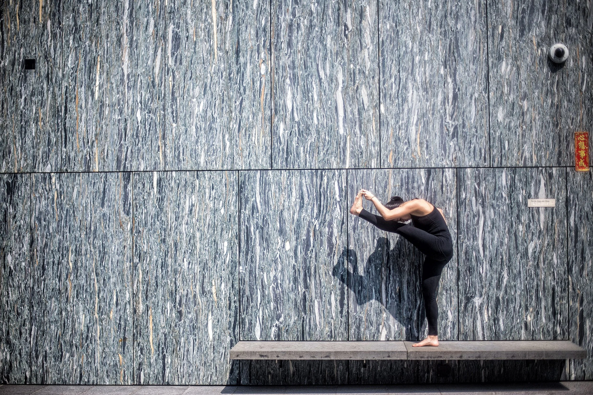 A person practicing yoga, standing on a platform and doing a forward bend stretch with their hands holding one foot behind their back, against a marble wall background.