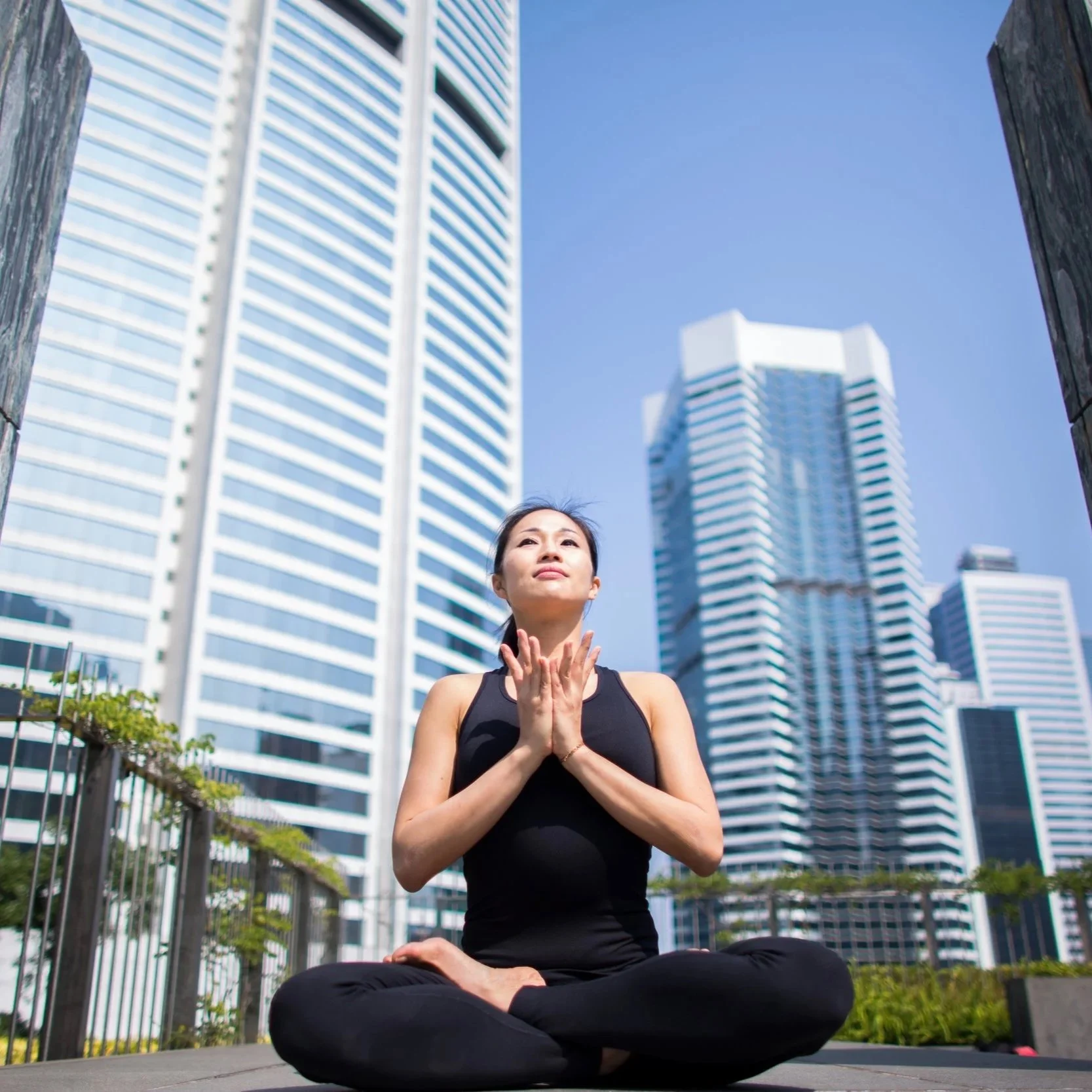A woman practicing yoga outdoors in a city, sitting cross-legged with hands in a prayer position against a backdrop of tall modern skyscrapers and blue sky.