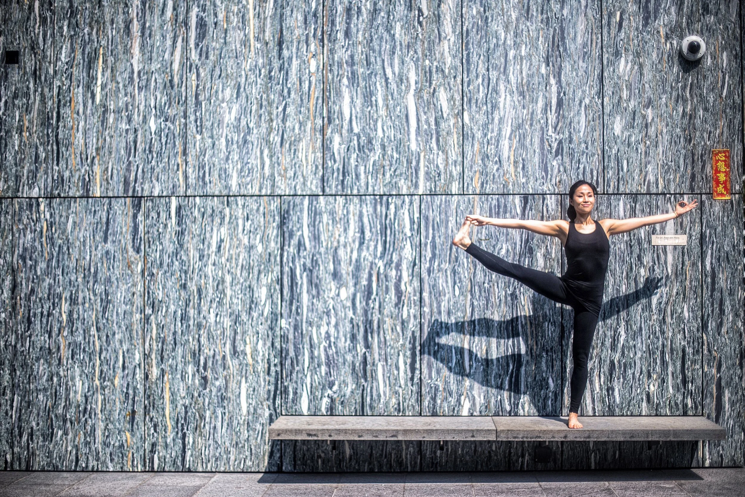 A woman in black athletic clothing practicing yoga on a ledge against a textured stone wall, standing on one leg with the other leg bent and held behind her while extending her arms outward.