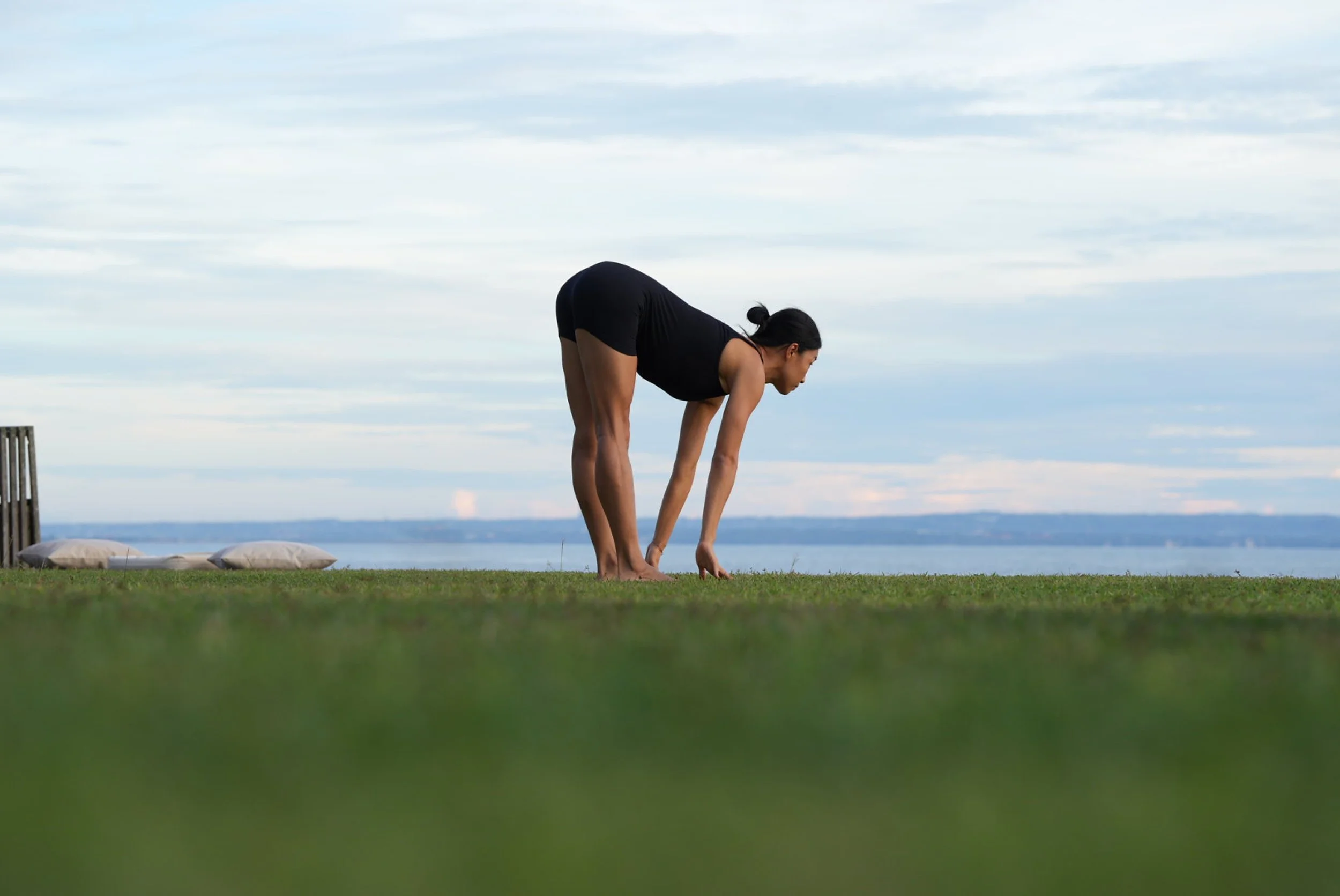 A woman practicing yoga outdoors on a grassy area, bending forward with her hands near her feet, with pillows placed nearby and a body of water in the background under a cloudy sky.