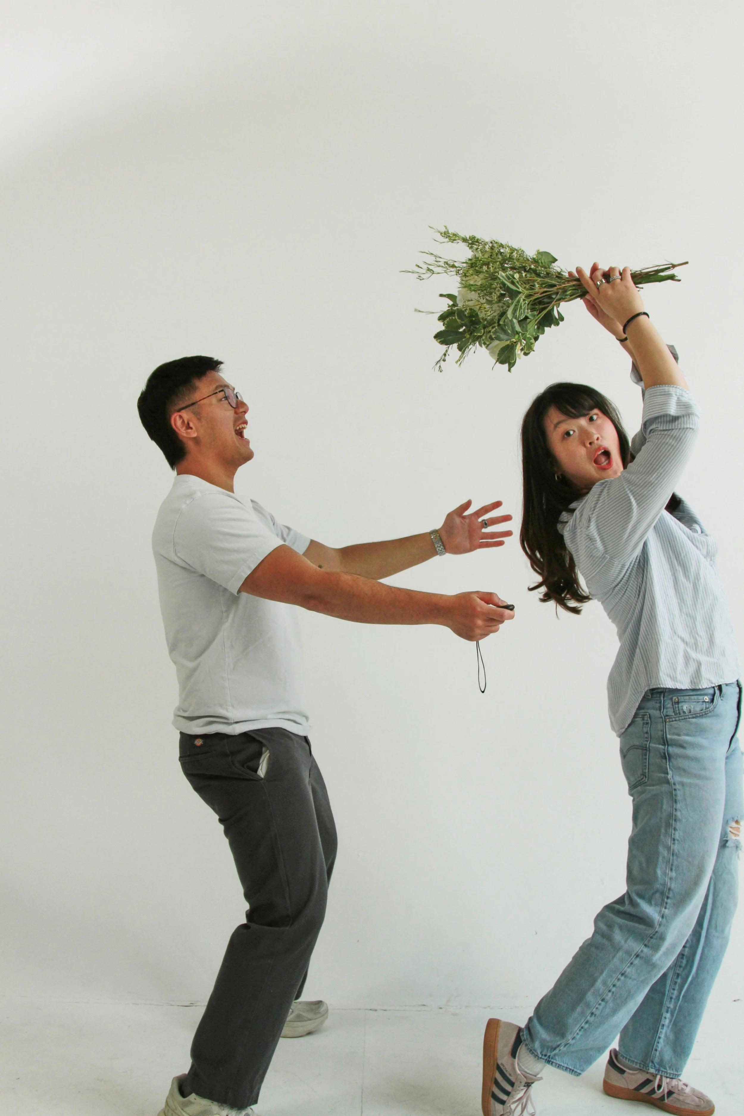 Two people, a man and a woman, appear to be playing or acting out a scene. The woman is holding a bunch of greenery and is leaning back with her mouth open, while the man is reaching out towards her with an excited expression. They are standing again