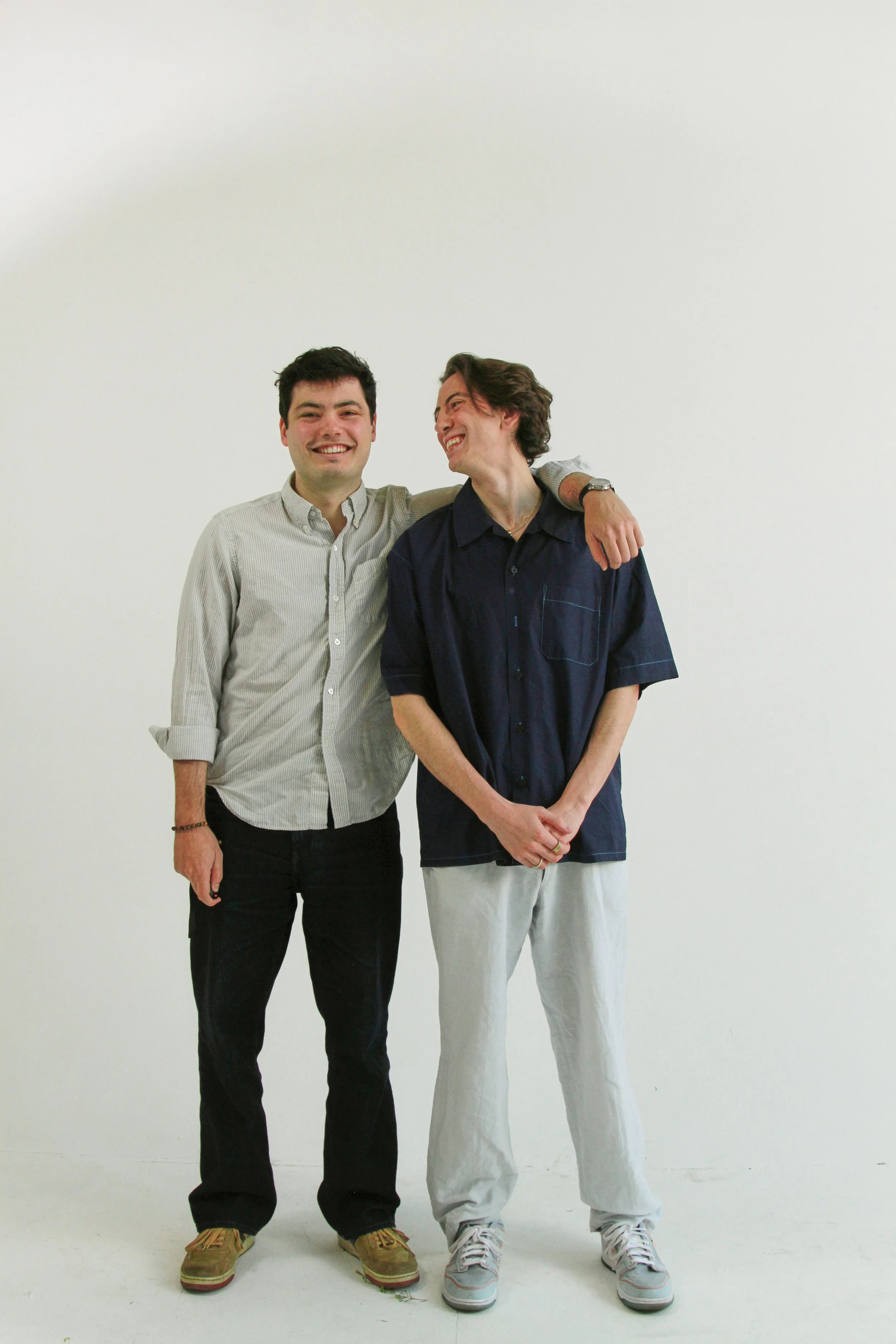 Two young men standing together against a plain white background, smiling and embracing each other. One is wearing a light striped shirt and dark pants, and the other is in a navy blue shirt and light-colored pants.
