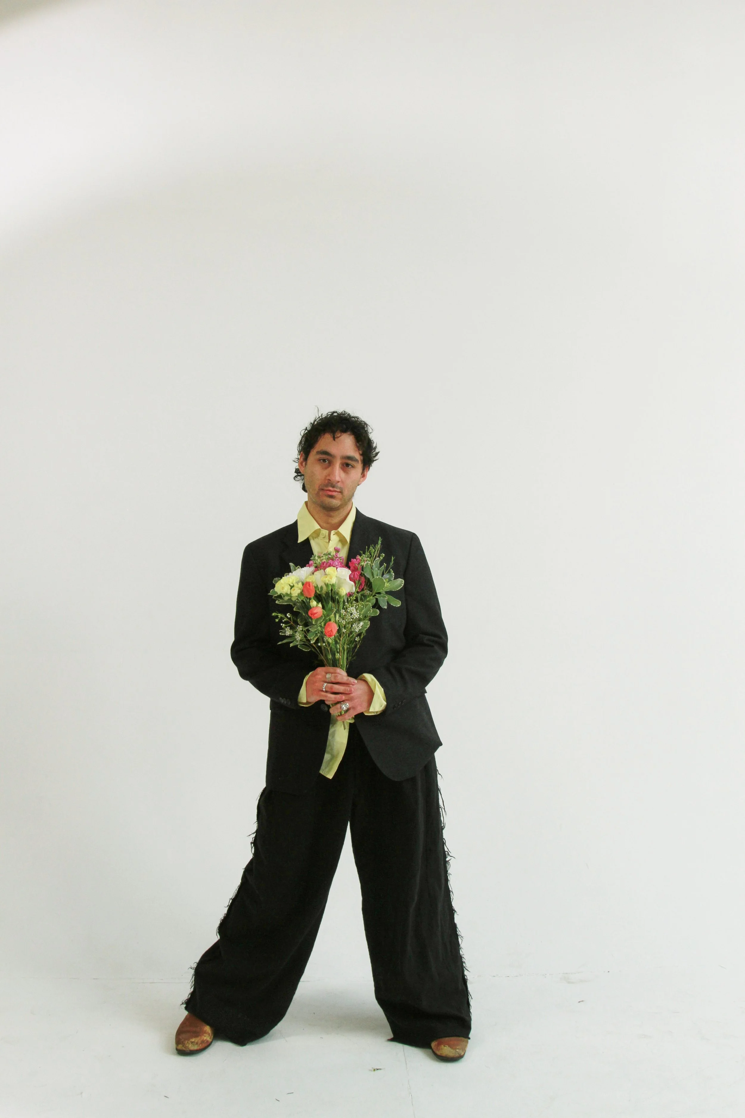 A man dressed in a black suit, yellow shirt, and beige shoes standing against a plain white background, holding a bouquet of flowers.