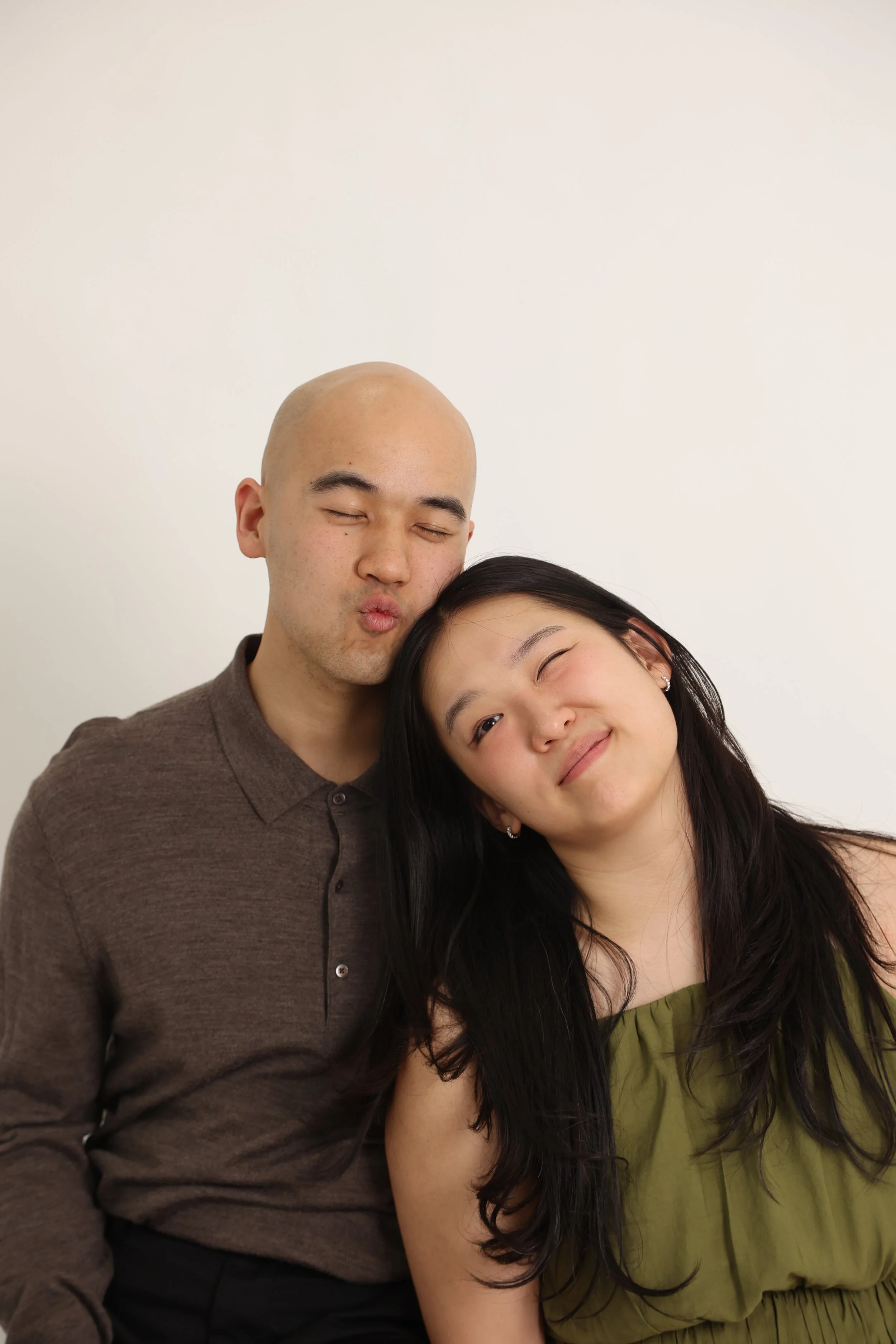 A man with a bald head and closed eyes making a kissing face next to a woman with long black hair smiling and leaning her head on his shoulder, both against a plain light background.