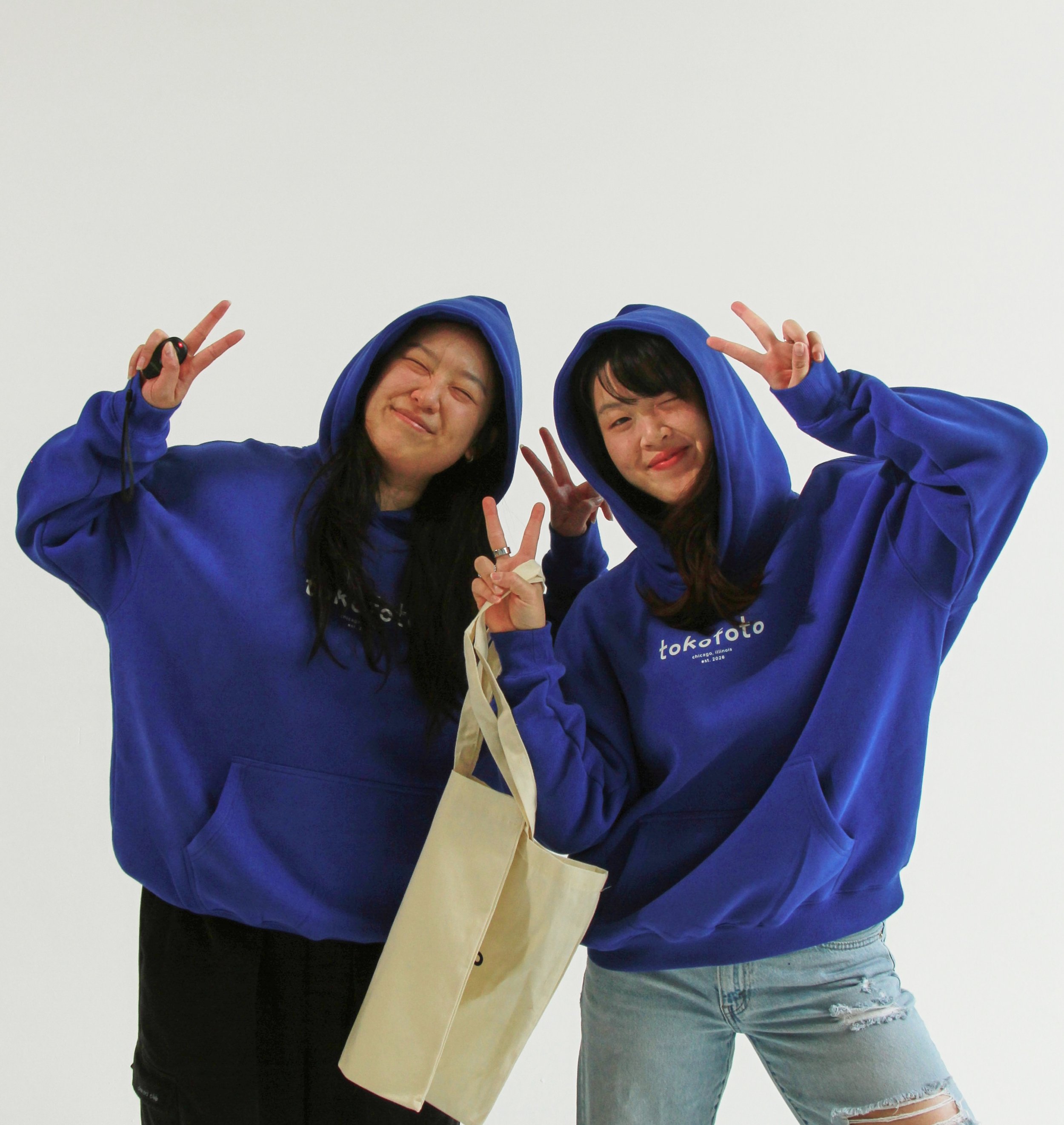Two women wearing blue hoodies making peace signs with their hands, smiling and posing against a plain white wall.