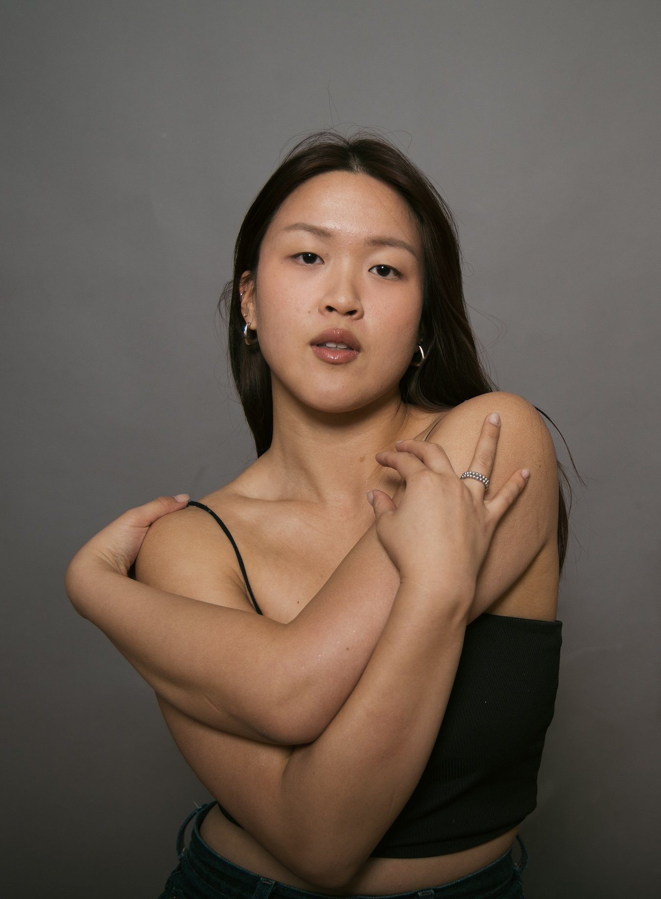 Portrait of a young woman with dark hair wearing earrings and a black spaghetti strap top, standing against a gray background, with her arms crossed over her chest.