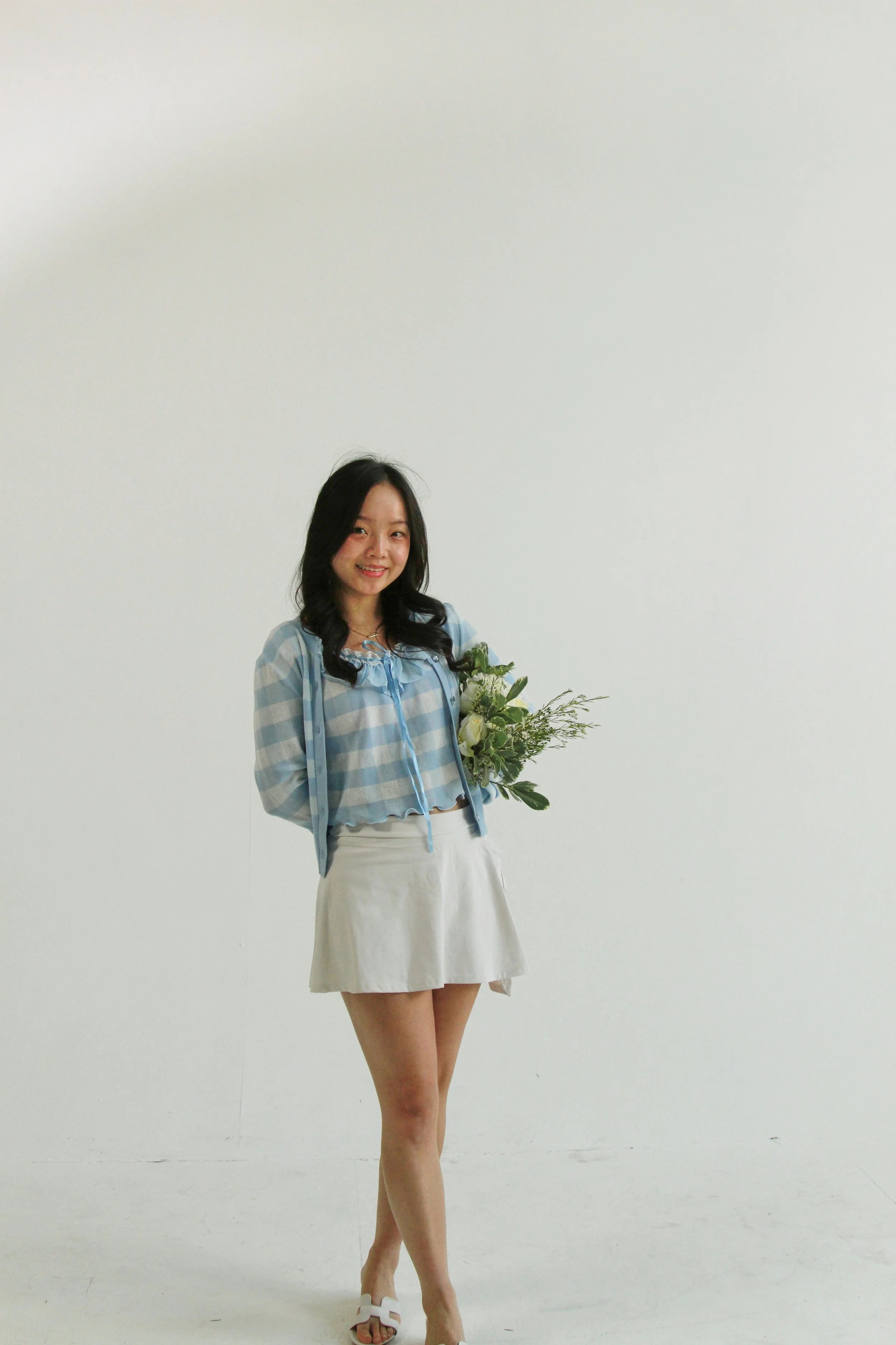 A young woman is standing against a white wall, holding a bouquet of white flowers with green foliage, and smiling at the camera. She is dressed in a blue and white checkered blouse, white skirt, and white shoes.