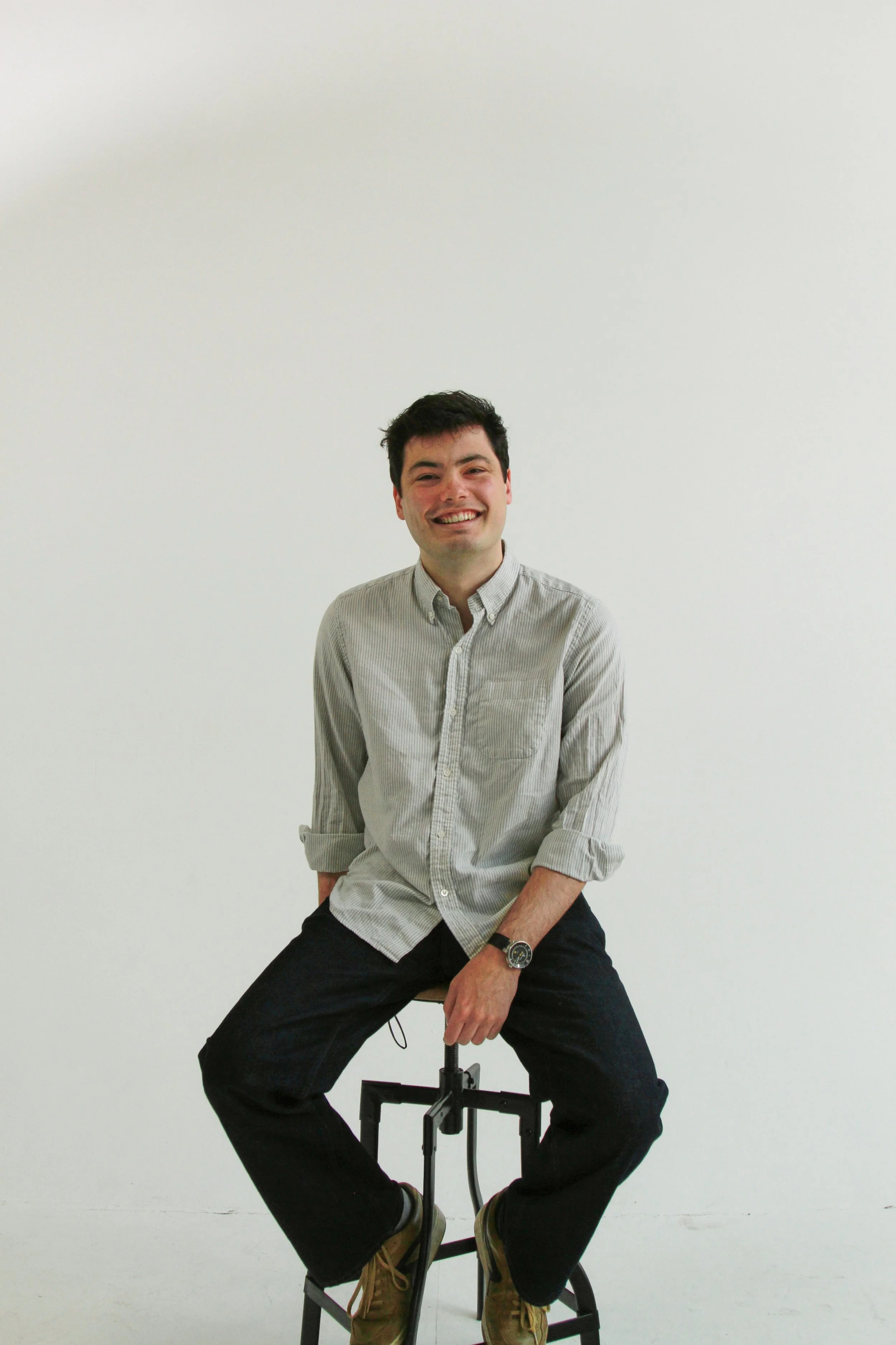 A young man with dark hair and a light-colored button-down shirt sitting on a stool against a plain white wall, smiling and looking at the camera.