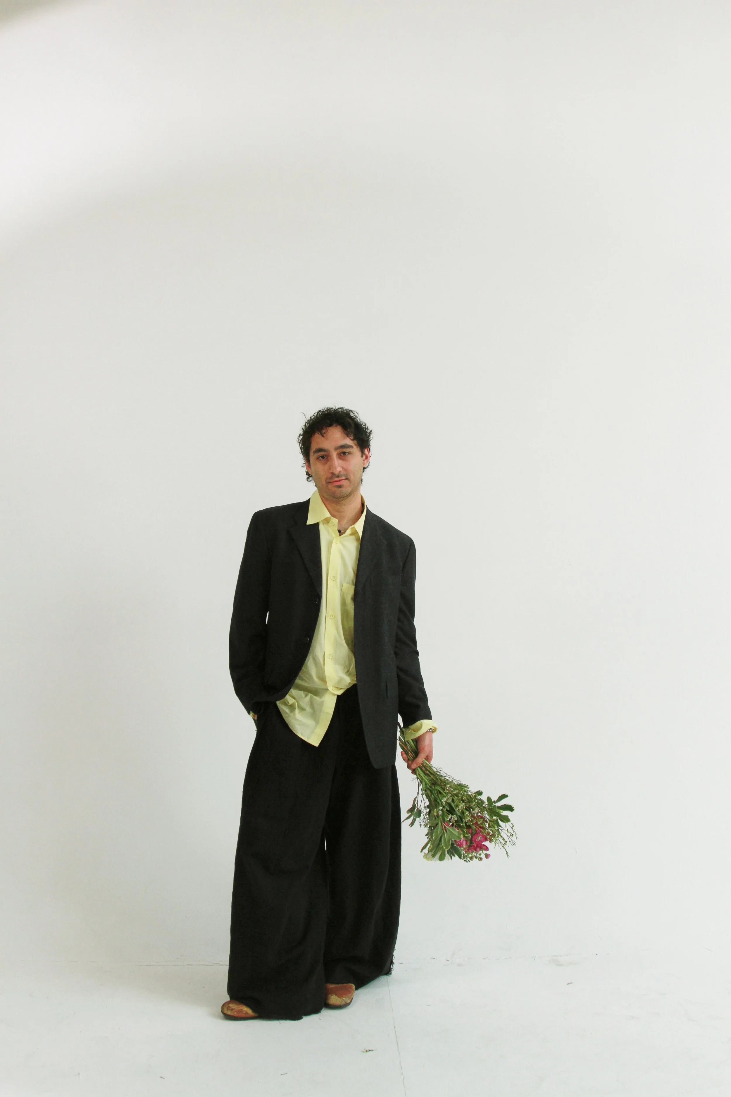 A man in a black suit with a yellow shirt holding a bouquet of pink and purple flowers stands against a plain white wall.