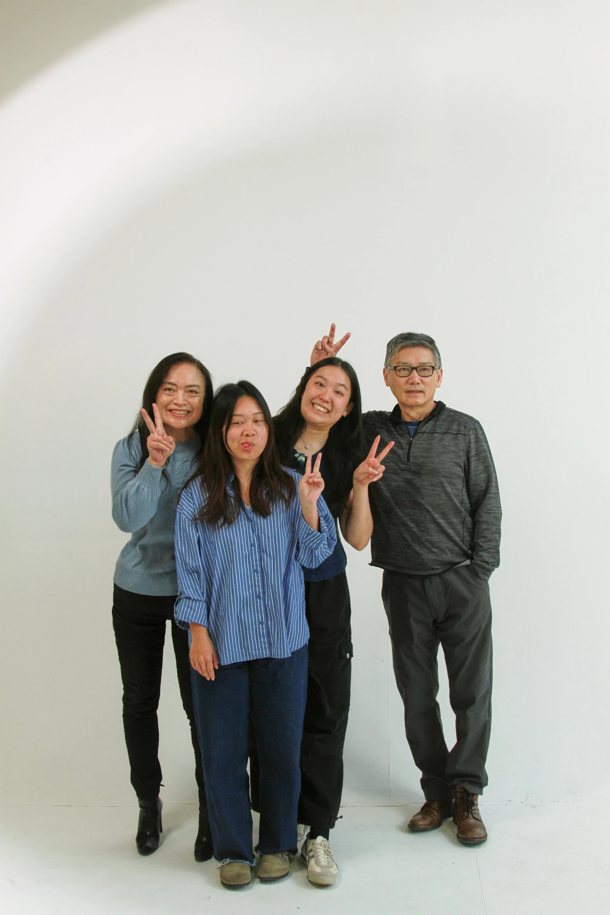 Group of five people posing together against a plain white wall, making peace signs and smiling at the camera.