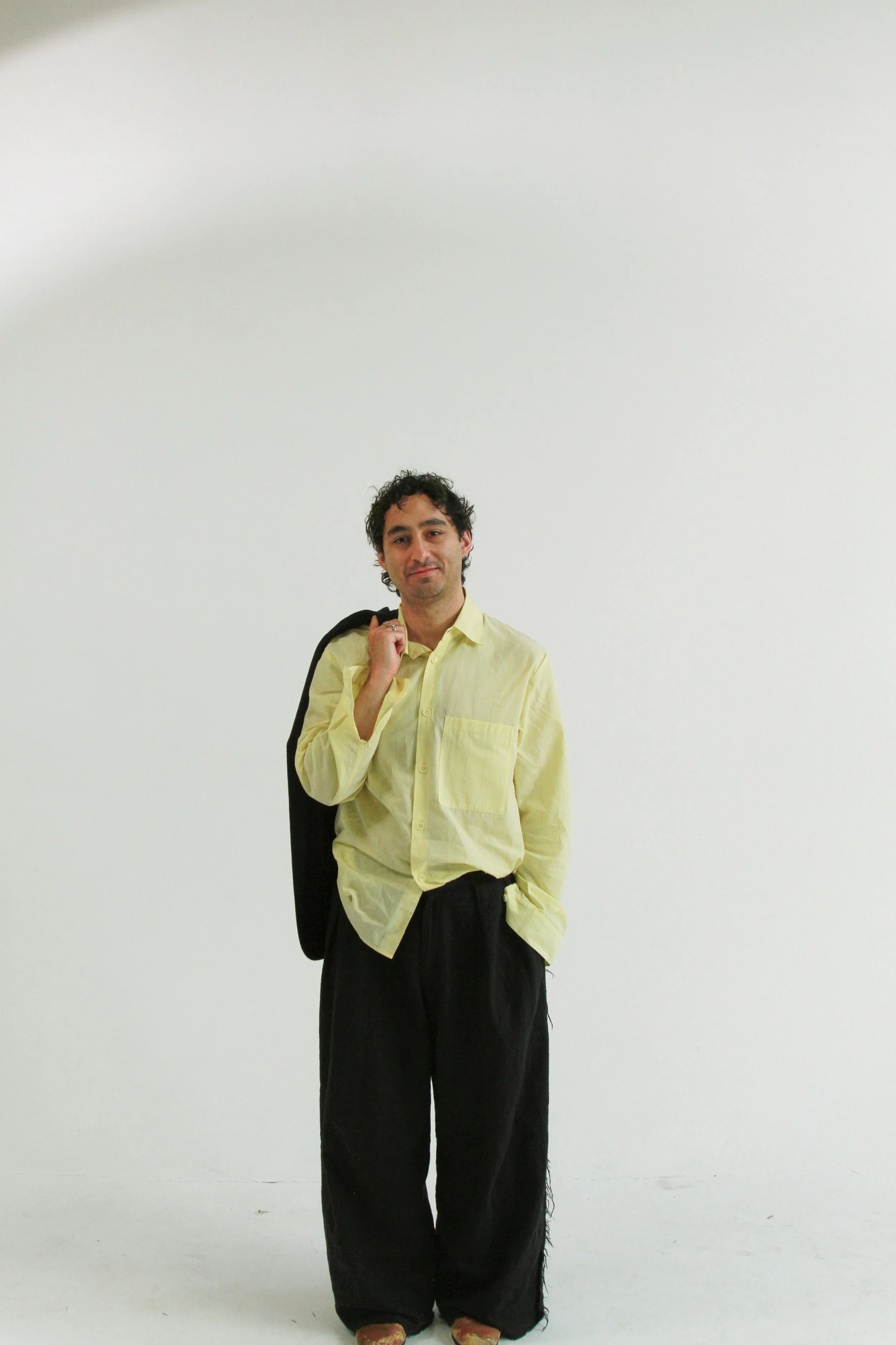 A man with dark curly hair, wearing a yellow button-down shirt and black pants, standing against a plain white background. He is carrying a black jacket over his right shoulder and is looking at the camera with a slight smile.
