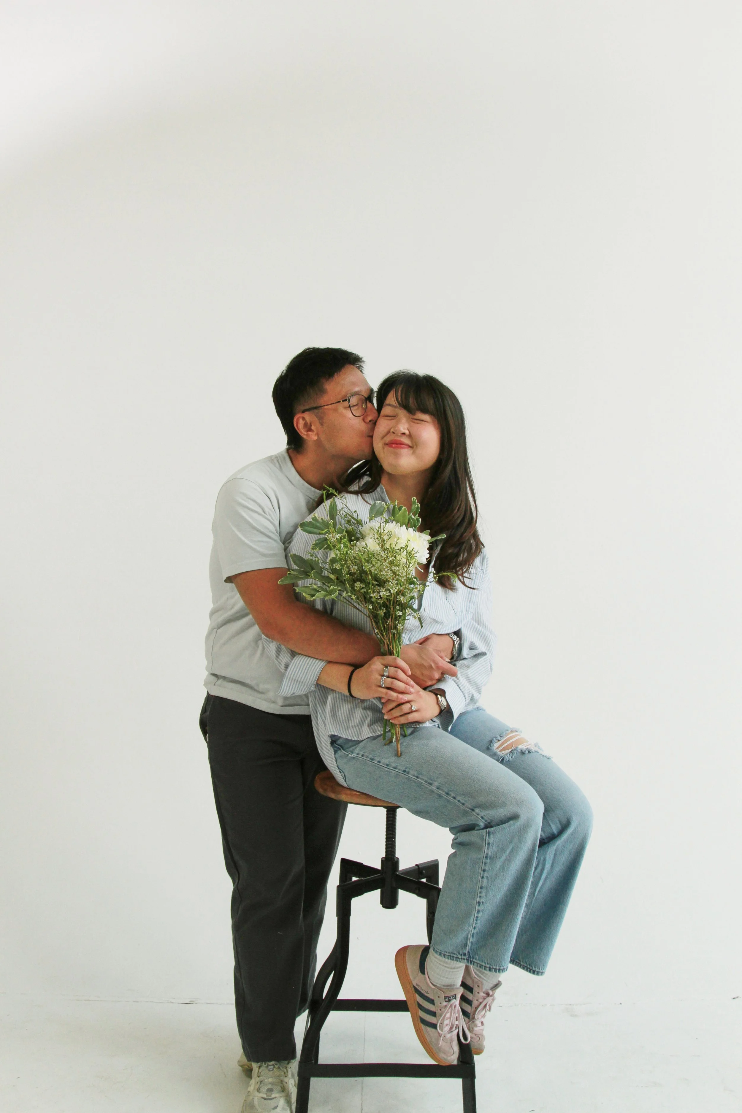 A man giving a woman a kiss on the cheek while she smiles with her eyes closed, sitting on a stool, holding a bouquet of flowers, with a plain white wall in the background.
