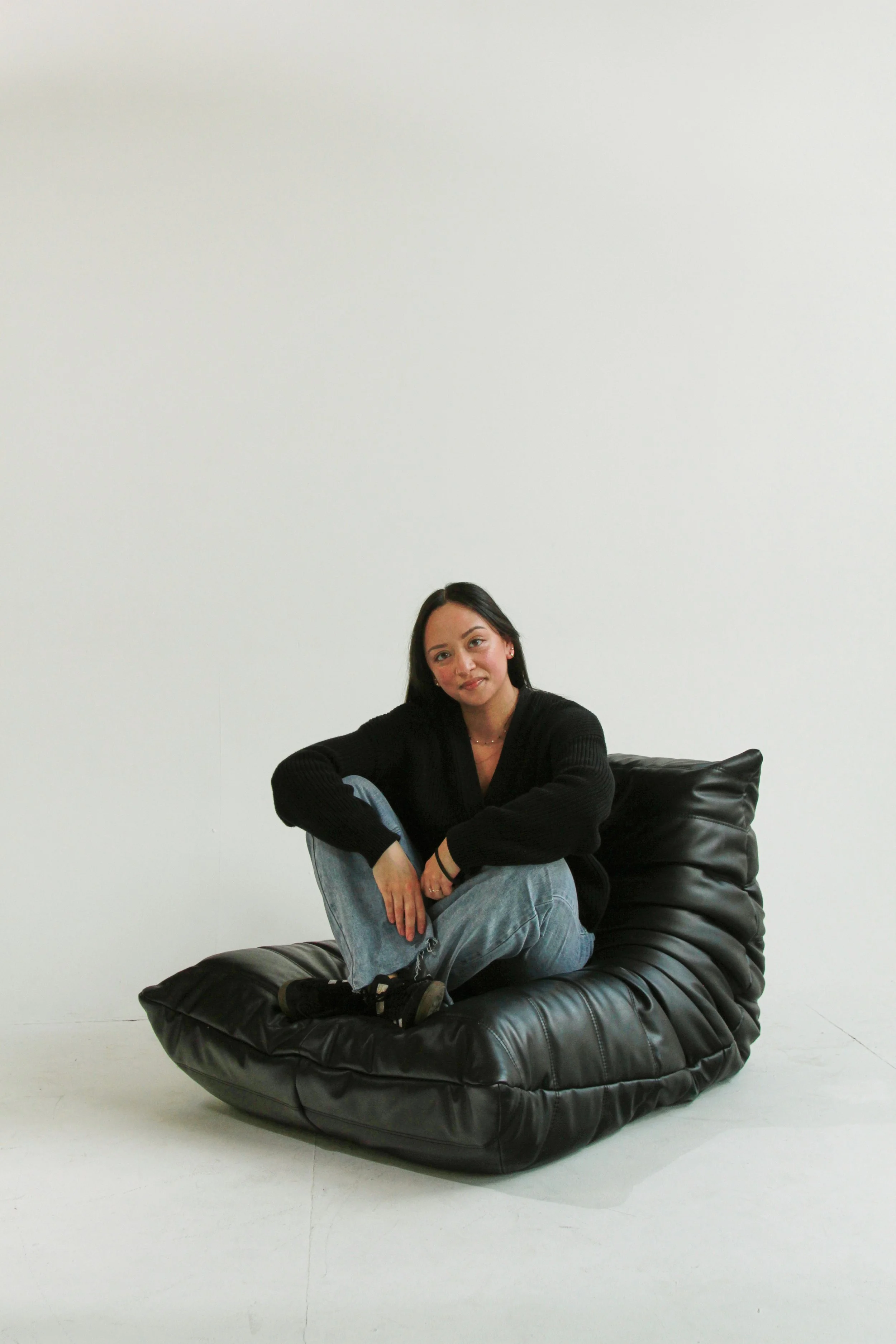 A woman sitting on a large black bean bag chair against a plain white wall, wearing a black sweater and jeans, with dark hair and a slight smile.
