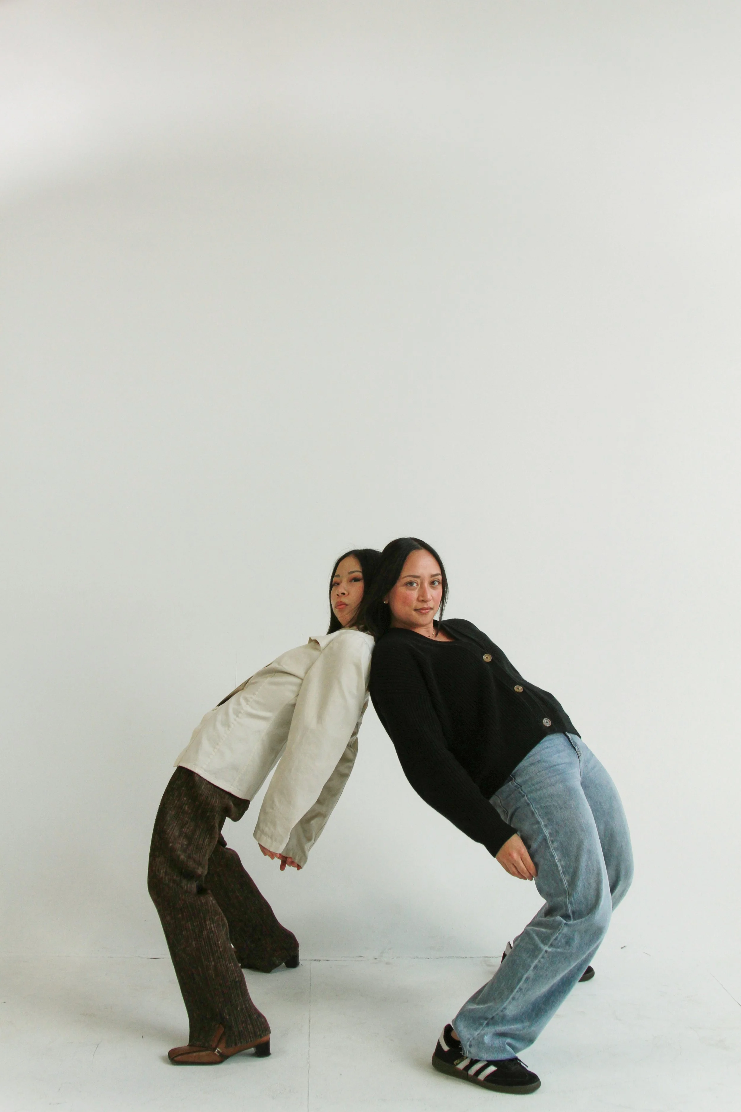 Two women standing against a plain white background, leaning back to back, holding hands, and looking at the camera.